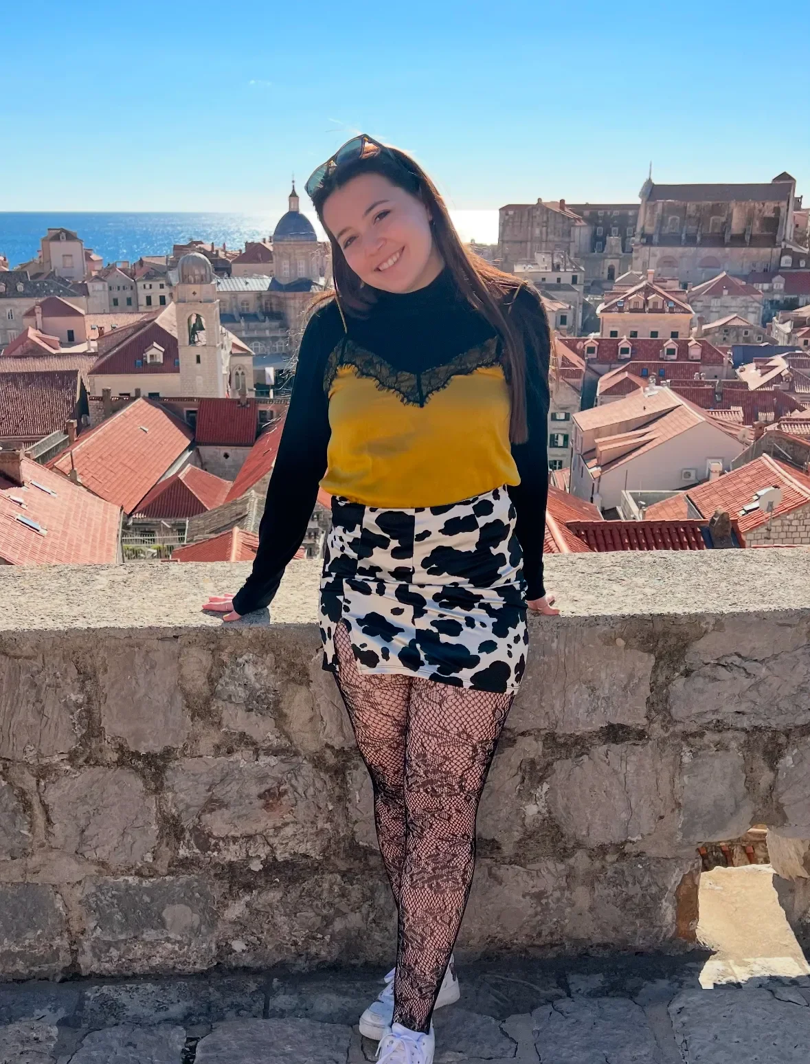 A young woman with long dark hair, wearing sunglasses, a yellow and black top, a black and white animal print skirt, fishnet tights, and white sneakers, standing outdoors on a stone ledge with a cityscape and the ocean in the background.