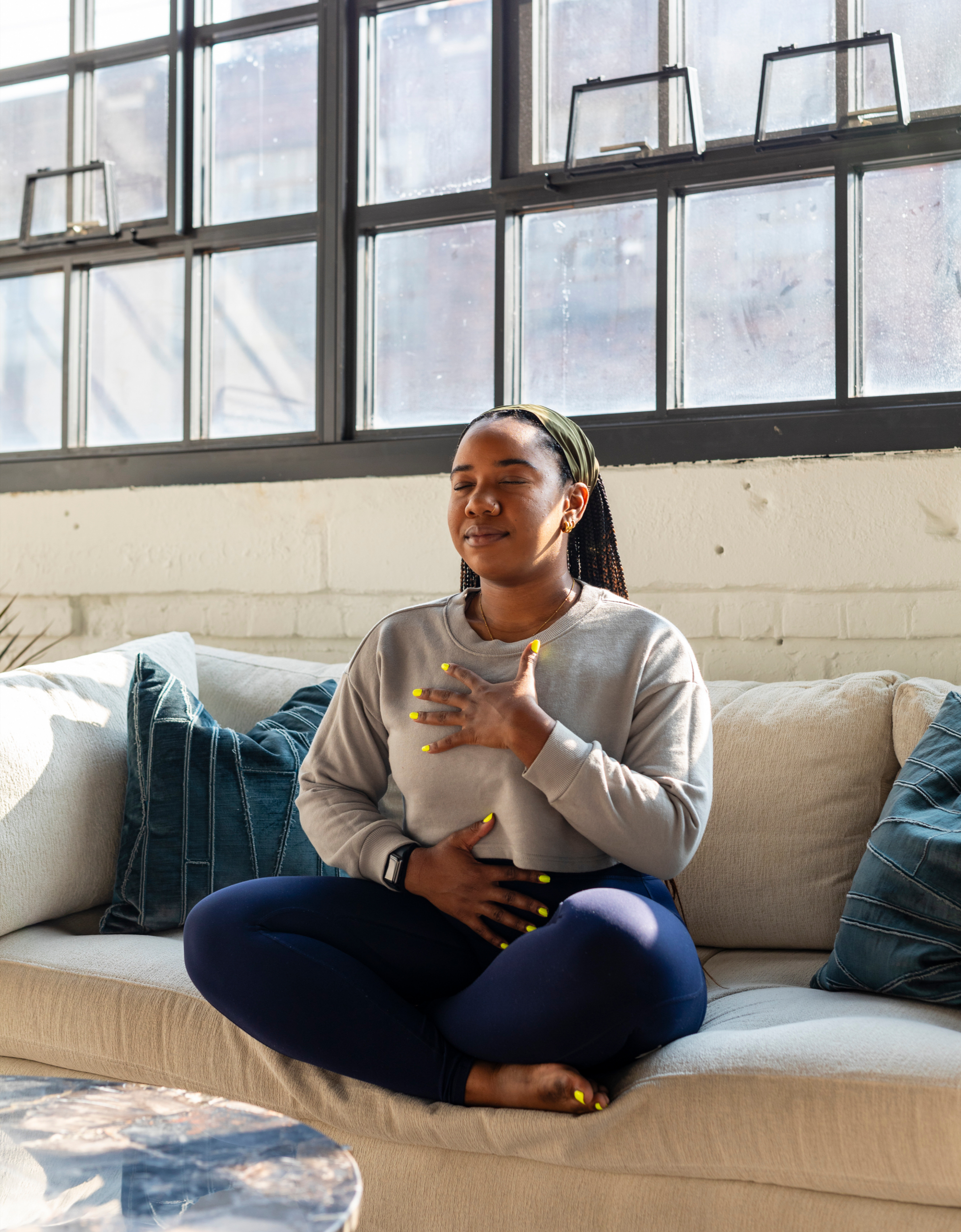 A woman sitting cross-legged on a beige sofa with her eyes closed, holding her hand to her chest and stomach, appearing to meditate or practice mindfulness in a well-lit room with large windows.