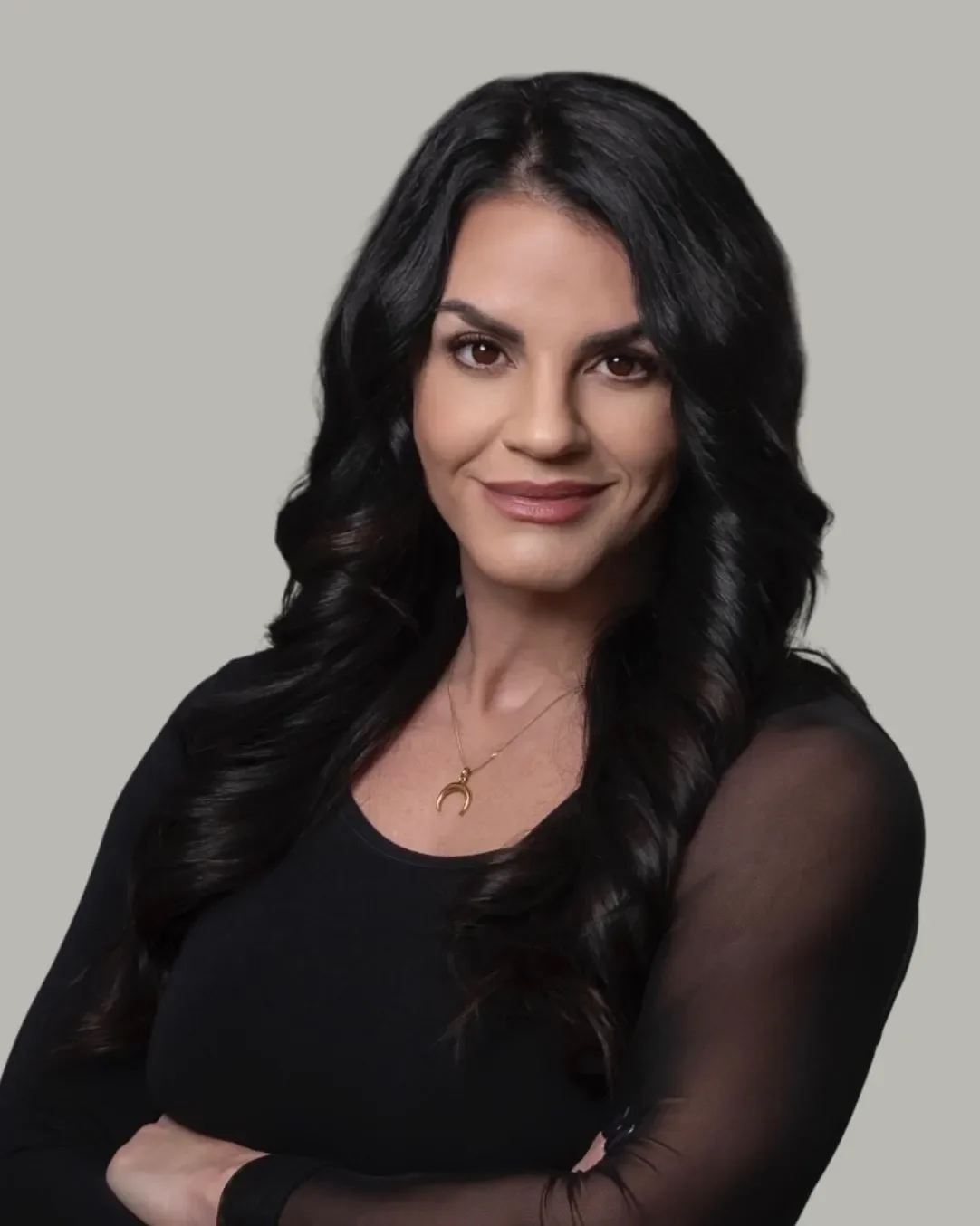 A woman with long, wavy black hair wearing a black top with sheer sleeves and a gold crescent moon necklace, smiling at the camera against a plain gray background.