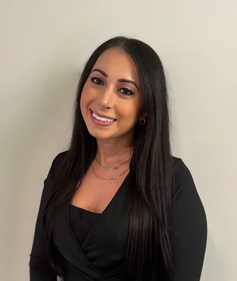 A smiling woman with long black hair and light makeup wearing a black top and layered gold necklaces, standing against a plain off-white background.
