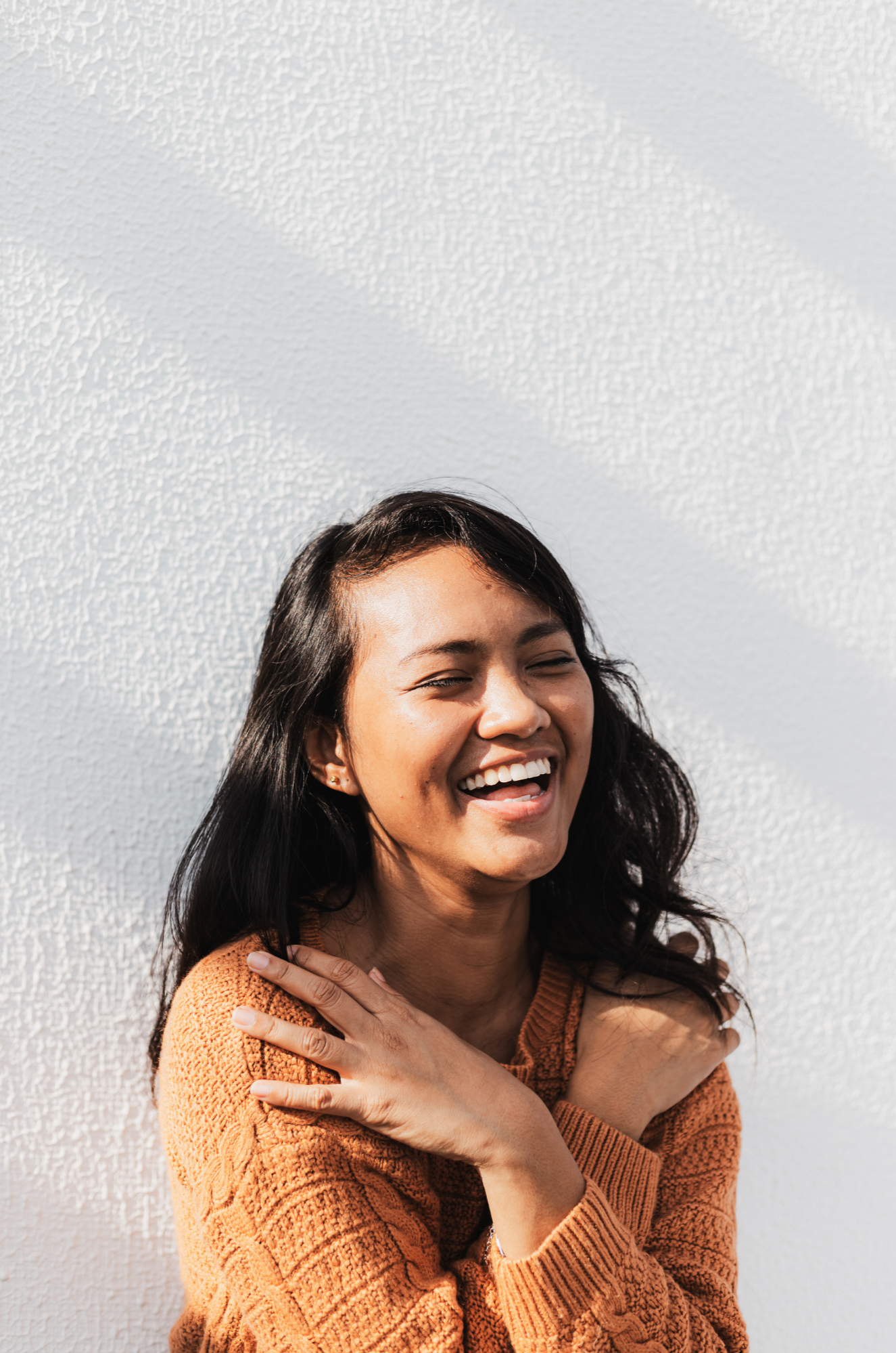 A woman with long dark hair and a bright smile, wearing an orange sweater, standing against a textured white wall.
