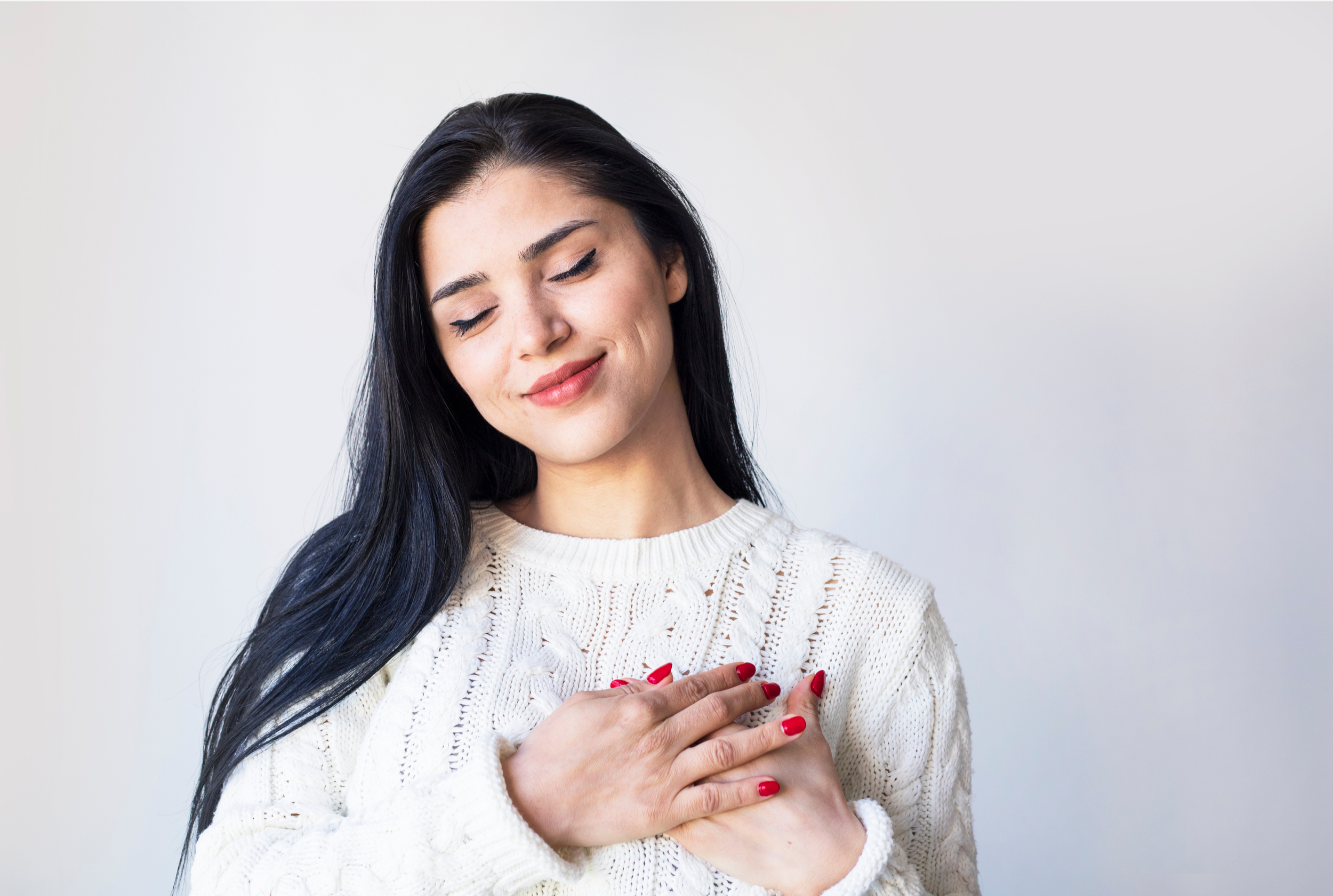 A woman with long dark hair wearing a white knit sweater, eyes closed, smiling gently, with her hand over her heart, standing against a plain white background.