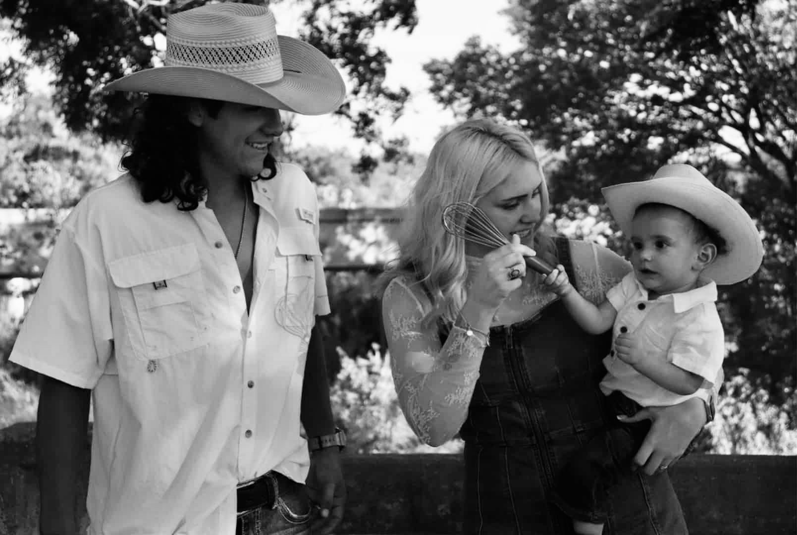 A woman holding a baby and a man standing nearby, all wearing cowboy hats, outdoors with trees in the background.