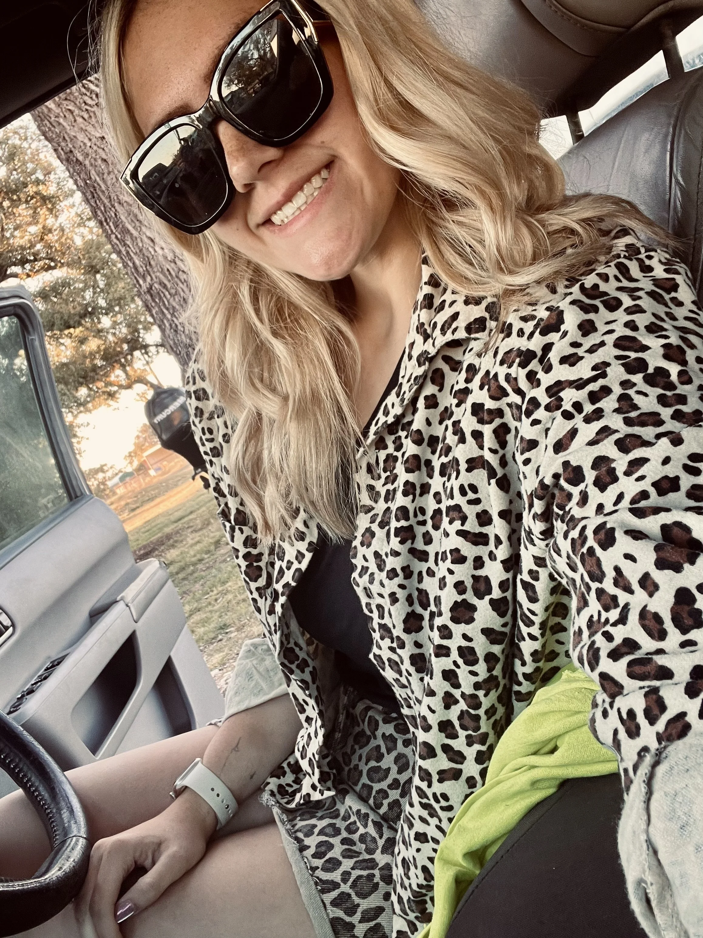 A woman with blonde, wavy hair wearing large black sunglasses, a leopard print shirt, and a black top. She is sitting in a vehicle, smiling, with sunlight and trees visible outside.