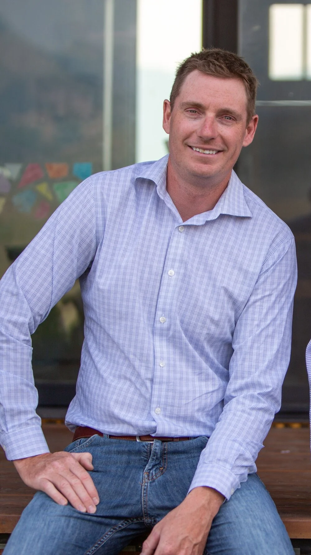 A man with short brown hair, wearing a light blue checkered shirt and jeans, sitting outdoors in front of a building with reflective windows.