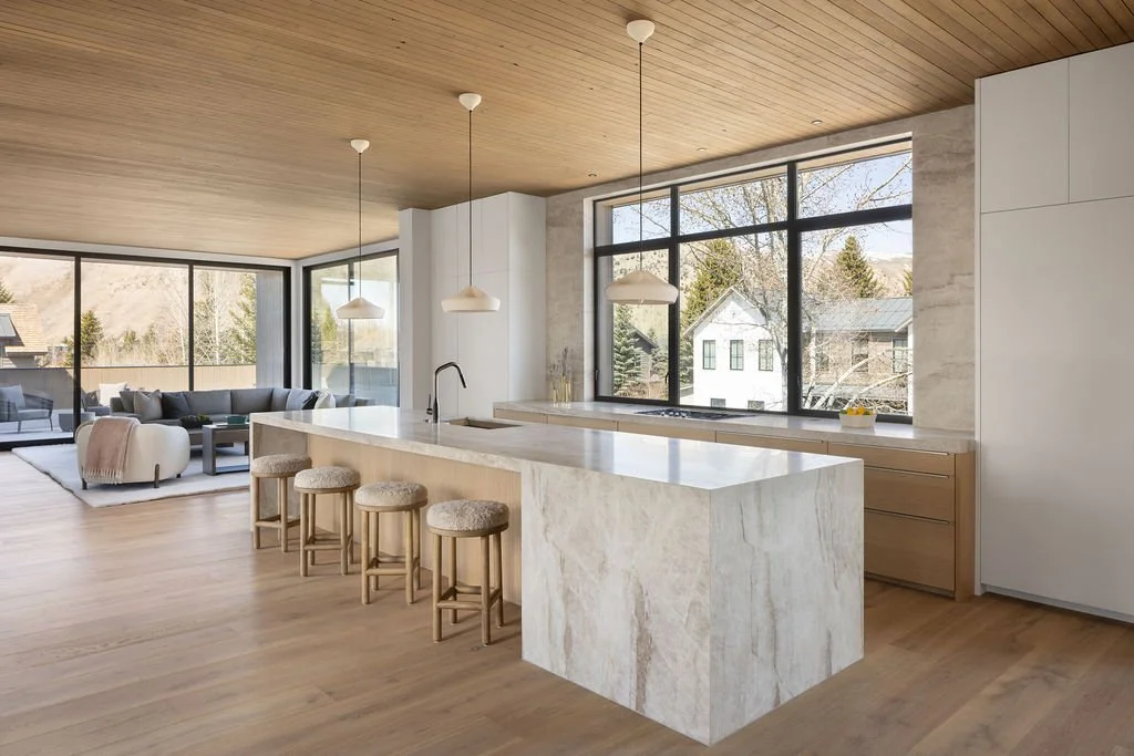 Modern kitchen with a large marble island, four bar stools, and three hanging pendant lights. Open-concept living area with large windows and a view of neighboring houses and trees.