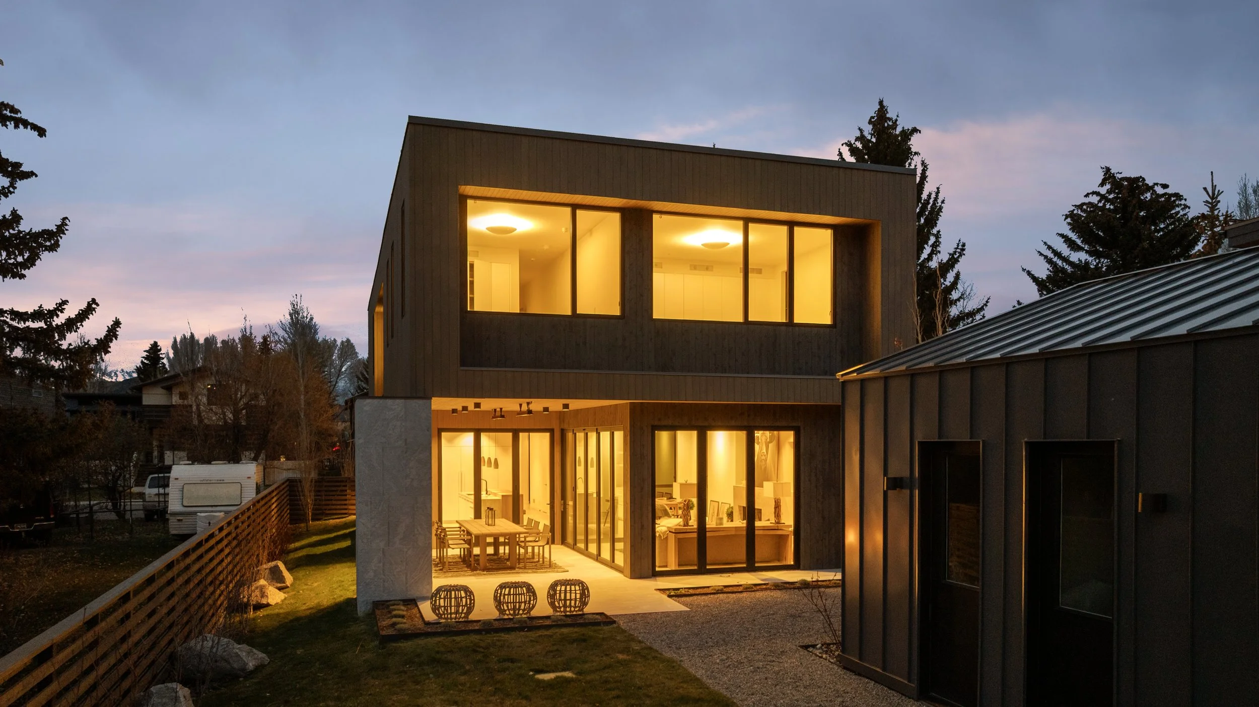 Modern two-story house with large illuminated windows at dusk, featuring a backyard with outdoor dining area and contemporary decor.