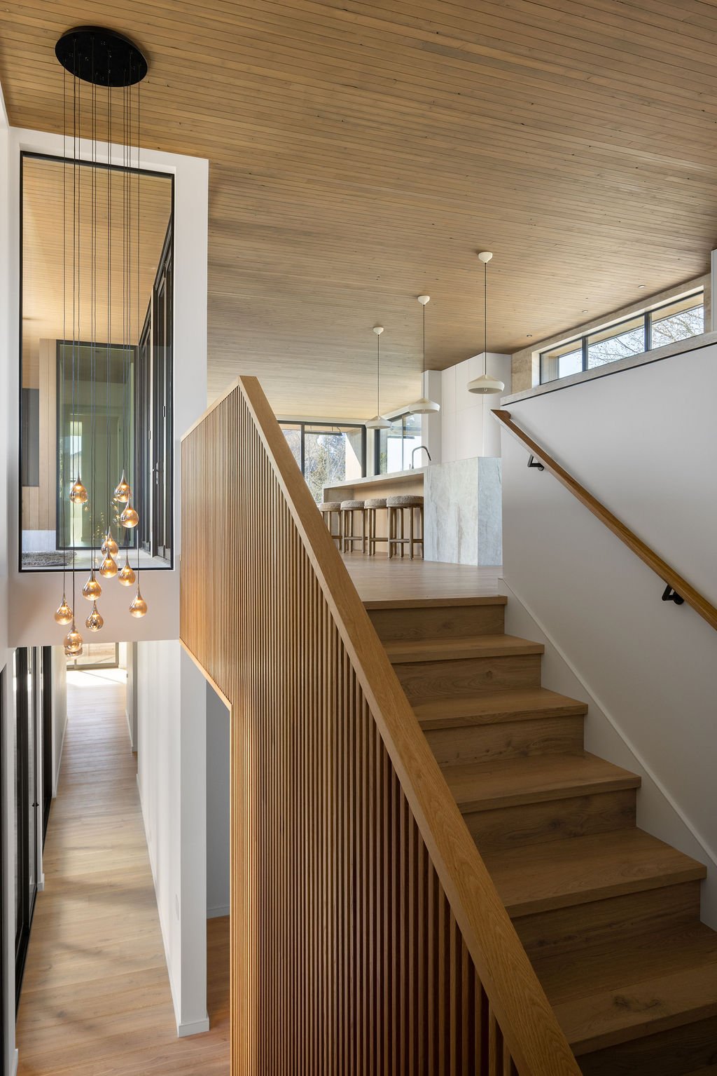 Interior view of a modern home featuring a staircase with wooden railing, hanging pendant lights, and a kitchen bar area with bar stools, large windows, and wooden ceiling.