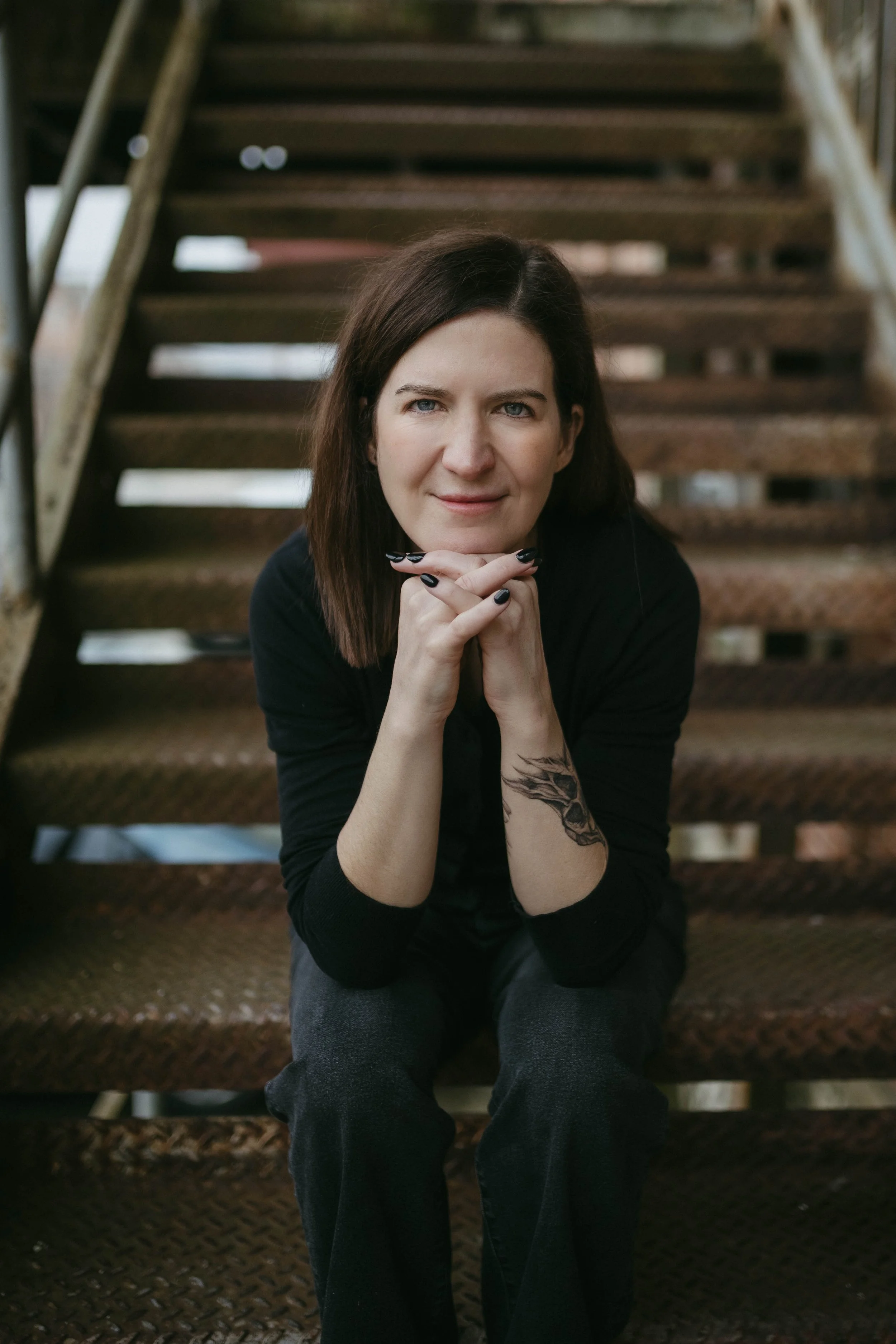 Woman in black shirt sits, hands folded under her chin, on rusty outdoor stairs.