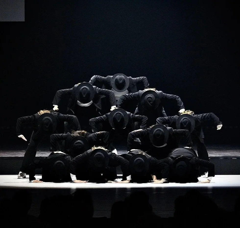 On Stage Dance competition - Twaalf dansers in zwart kostuum en hoed, in een choreografische opstelling op een podium met een zwarte achtergrond.