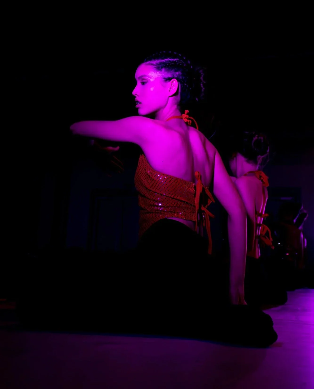 Vrouw danst in een donkere kamer onder roze neonlicht, met haar rug naar de camera, haar haar in vlechten, gekleed in een rood, gespiegelde top en zwarte broek, met een tweede persoon in de achtergrond.