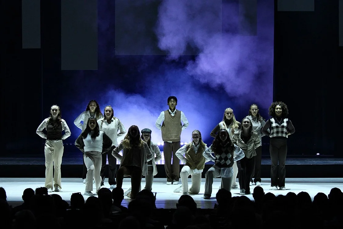 On Stage Dance competition - Een groep van twaalf dansers staat op een podium, met een duistere achtergrond en blauwe mist. Ze poseren met handen in de zij, gekleed in casual en sportieve kleding. Er is een publiek te zien op de voorgrond.