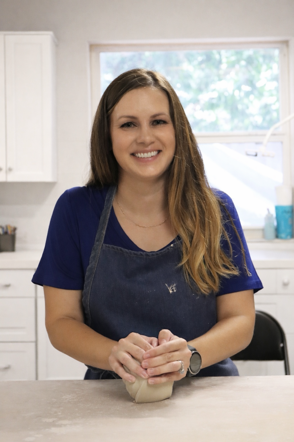 A woman in a blue shirt and apron smiling and kneading dough on a kitchen countertop.