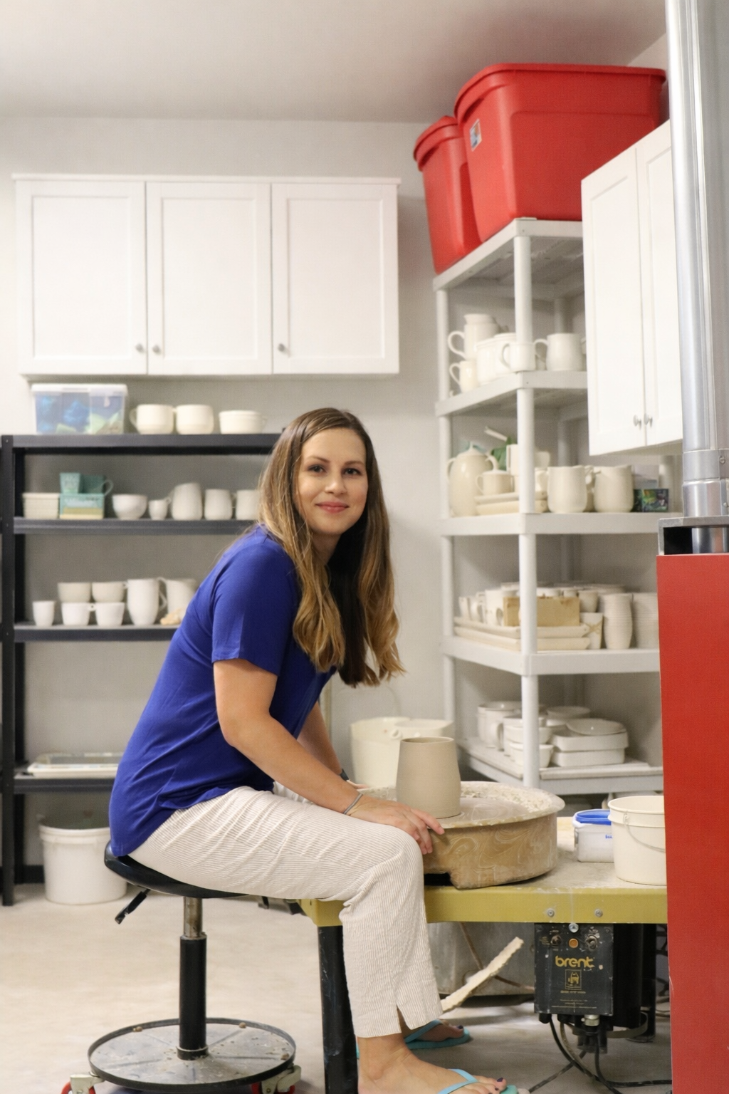 A woman with long brown hair, wearing a blue shirt and white pants, sits on a stool in a pottery studio, working on a ceramic piece on a spinning pottery wheel.