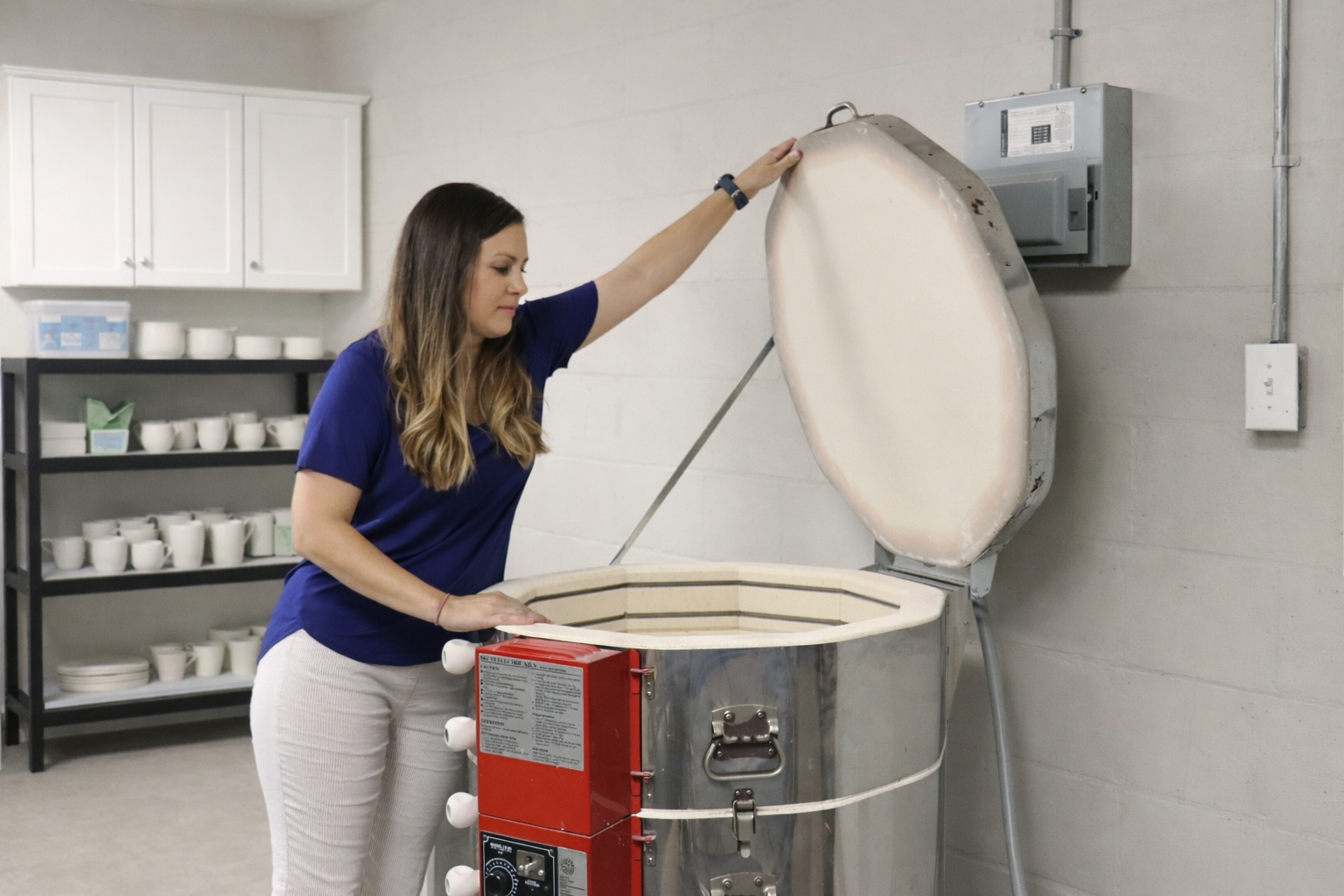 A woman opening a large, industrial microwave oven in a kitchen or ceramic studio.