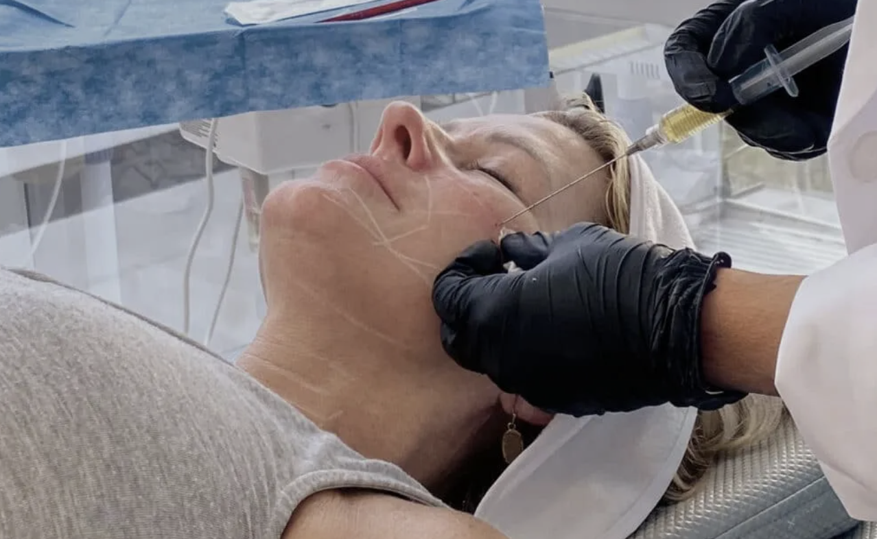 A woman receiving a cosmetic or medical injection in her face from a practitioner wearing black gloves, in a clinical setting.
