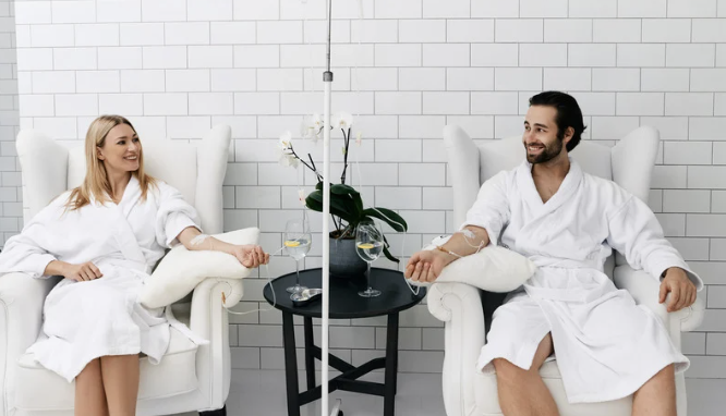 A woman and a man sitting in white armchairs, receiving IV therapy, in a spa or clinic setting with white tiled walls and a small black table with a flower vase.