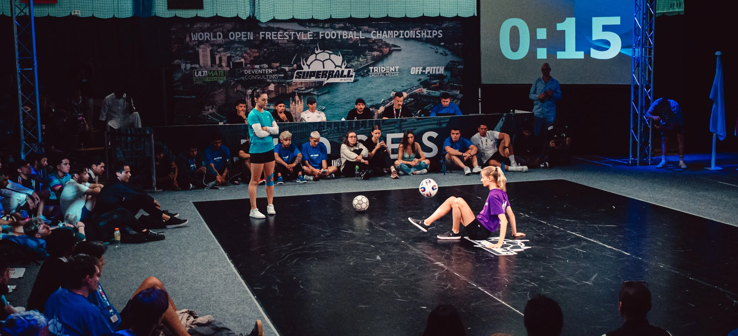 Zwei Frauen beim Fußballshow auf einer Bühne, eine sitzt am Boden und jongliert mit einem Ball, die andere steht. Ein Publikum sitzt um die Bühne. Im Hintergrund ein Bildschirm mit einer Timer-Anzeige und dem Text »World Open Freestyle Football Championships.«