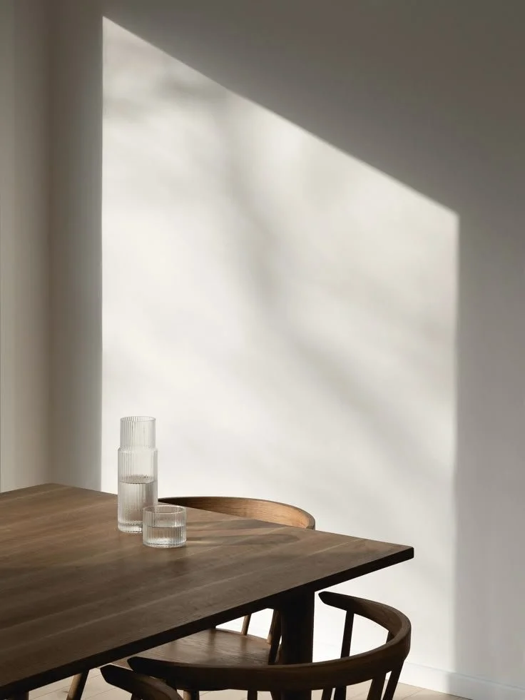 A wooden dining table with two glass water pitchers on it, surrounded by wooden chairs, with natural sunlight casting shadows on the wall.