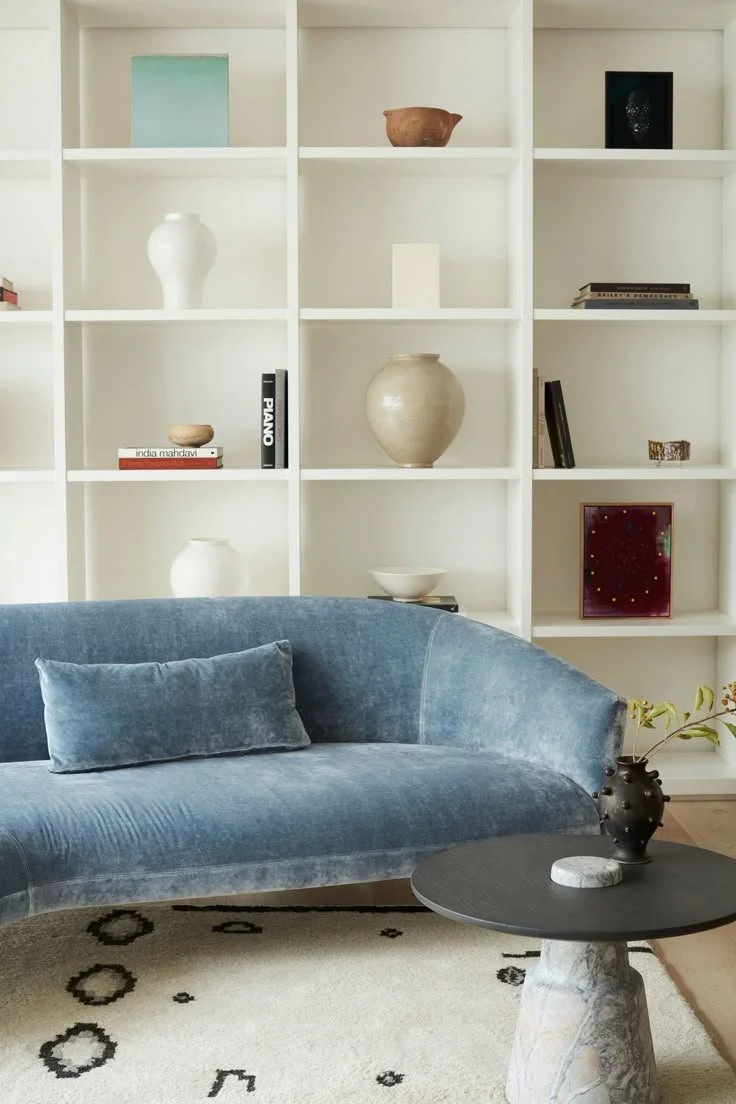Living room with a blue velvet sofa, a black circular side table with a vase, and a white built-in bookshelf with decorative items and books.
