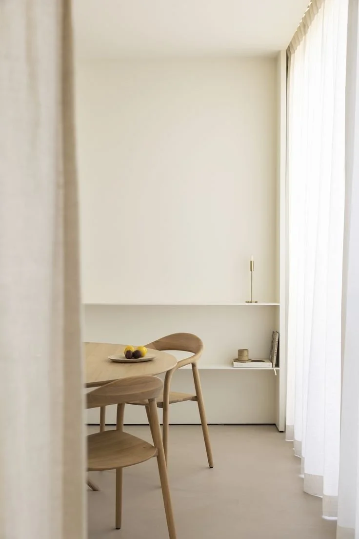 Minimalist dining area with a wooden table and chairs, a narrow white shelf with decorative items, and sheer curtains letting in natural light.