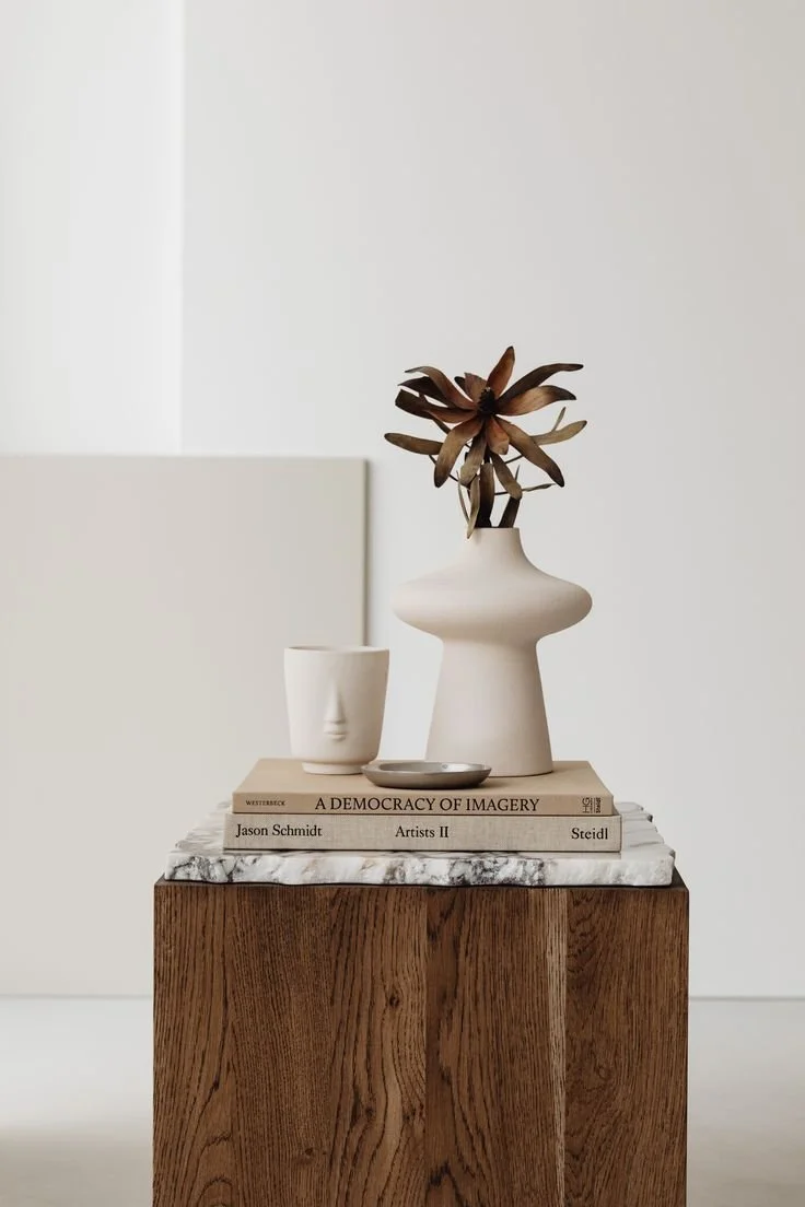 A still life of decorative objects on a wooden pedestal, including a white vase with a dried plant, a white face-shaped cup, a small silver dish, and stacked books against a neutral background.