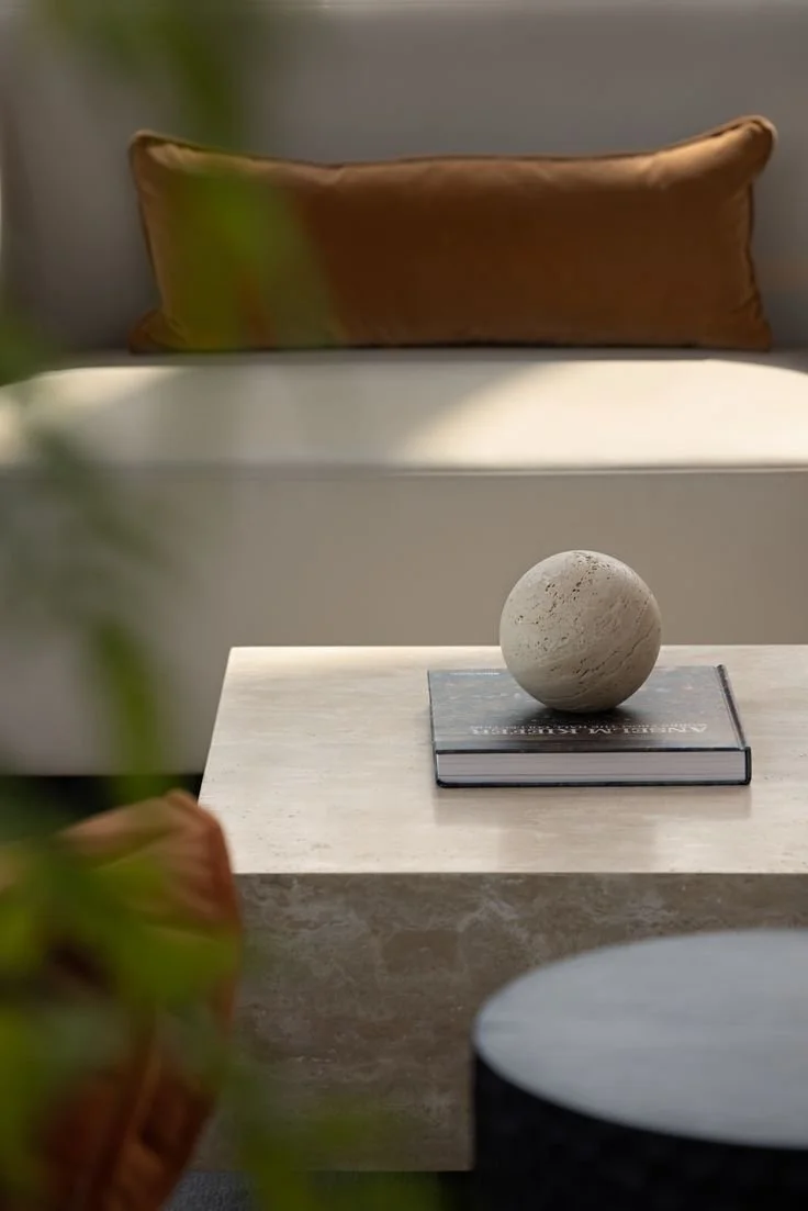 Close-up of a white marble table with a spherical stone sculpture resting on a closed book in a modern living room setting. A beige pillow is visible in the background.