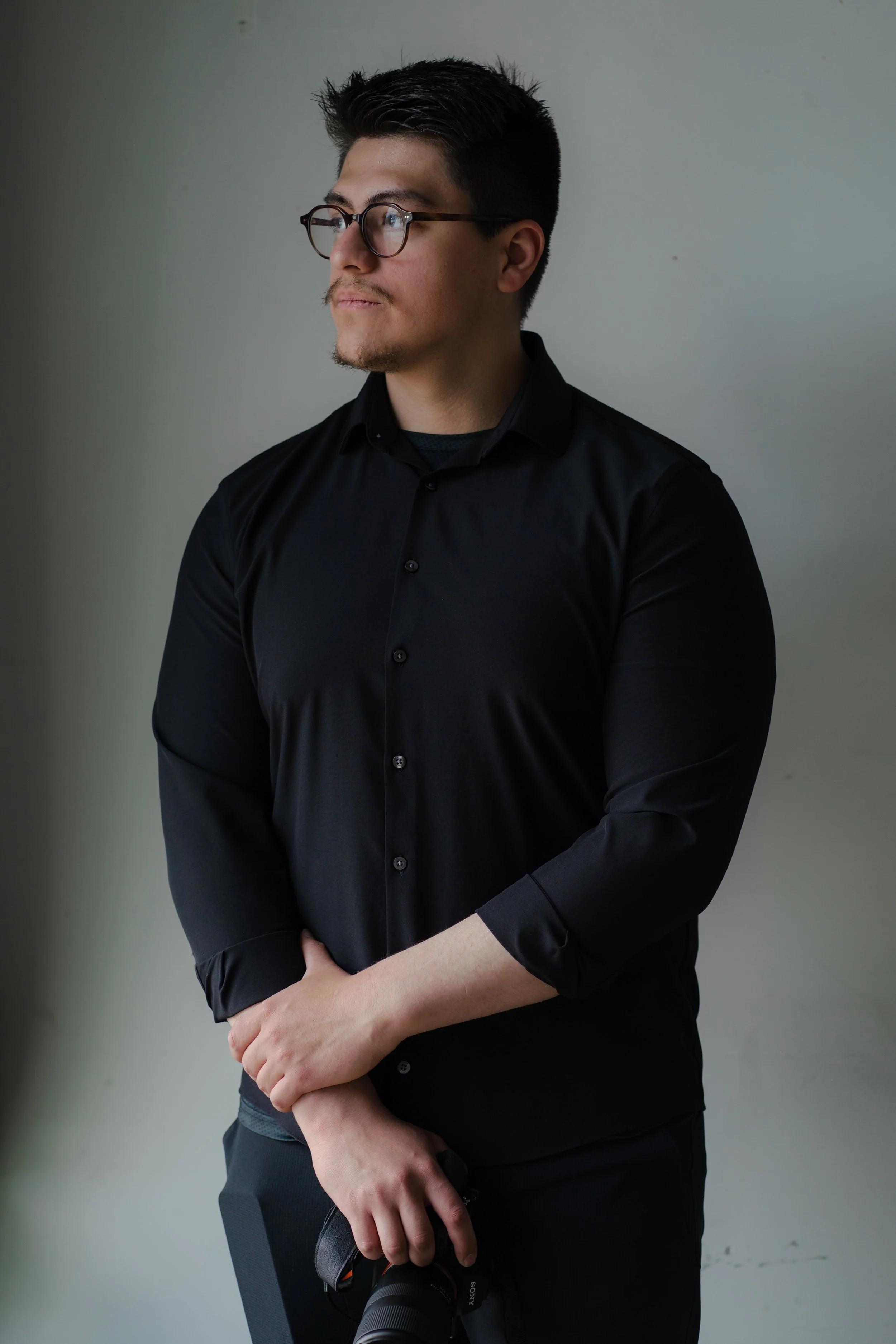 A man with glasses wearing a black shirt holding a camera, standing against a light gray wall.