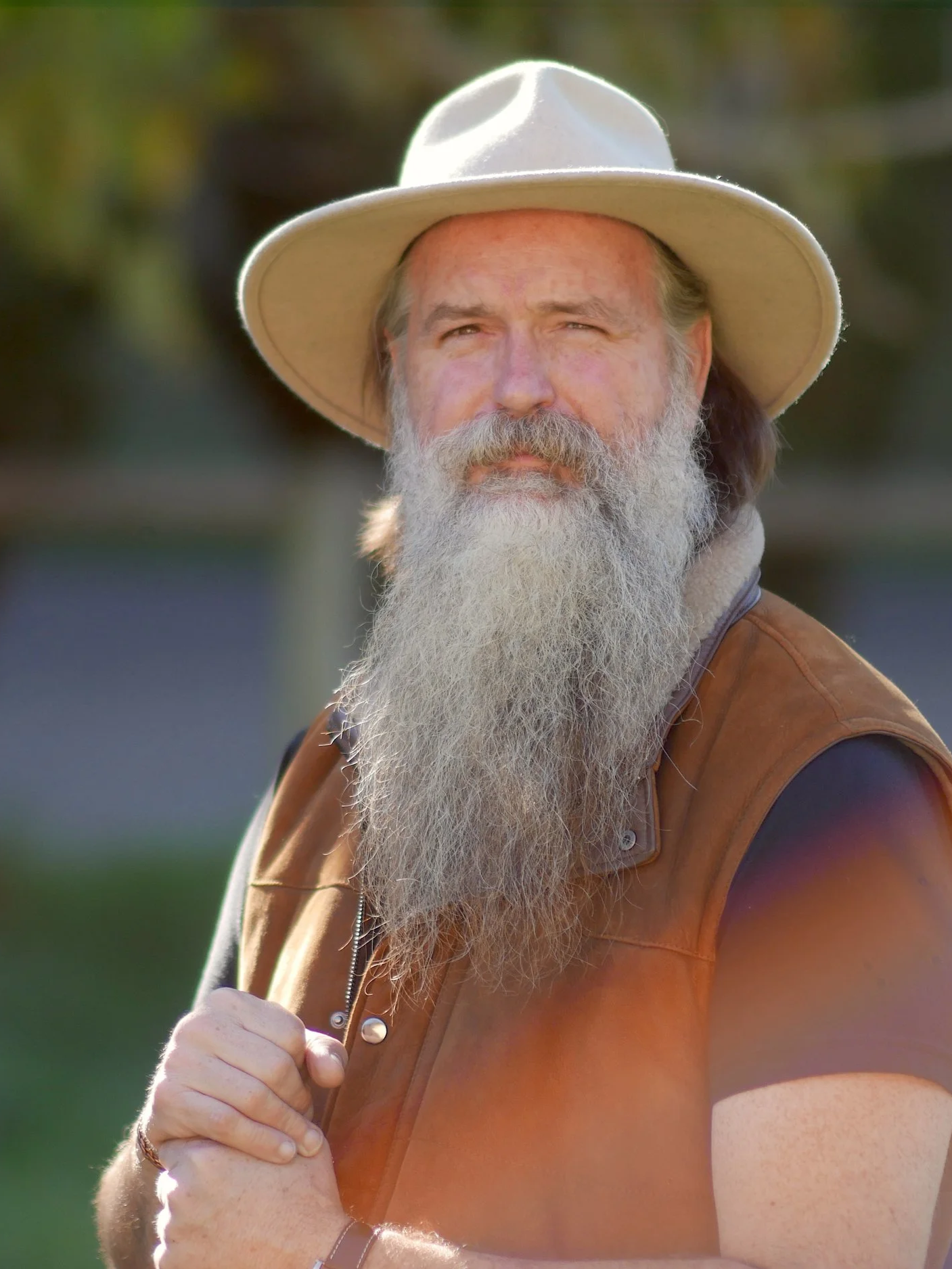 A bearded man wearing a wide-brimmed hat and brown vest, standing outdoors with a blurry background.
