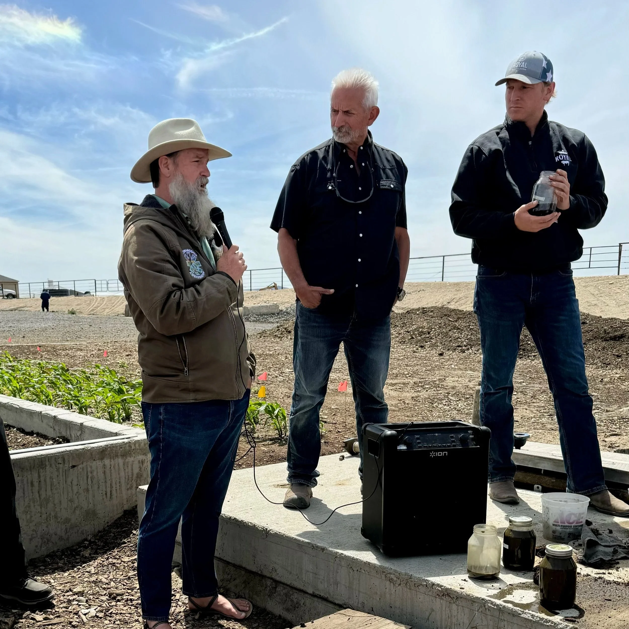 Three men standing outdoors near a raised garden bed with jars of dark liquids, listening to one man speak into a microphone under a partly cloudy sky.