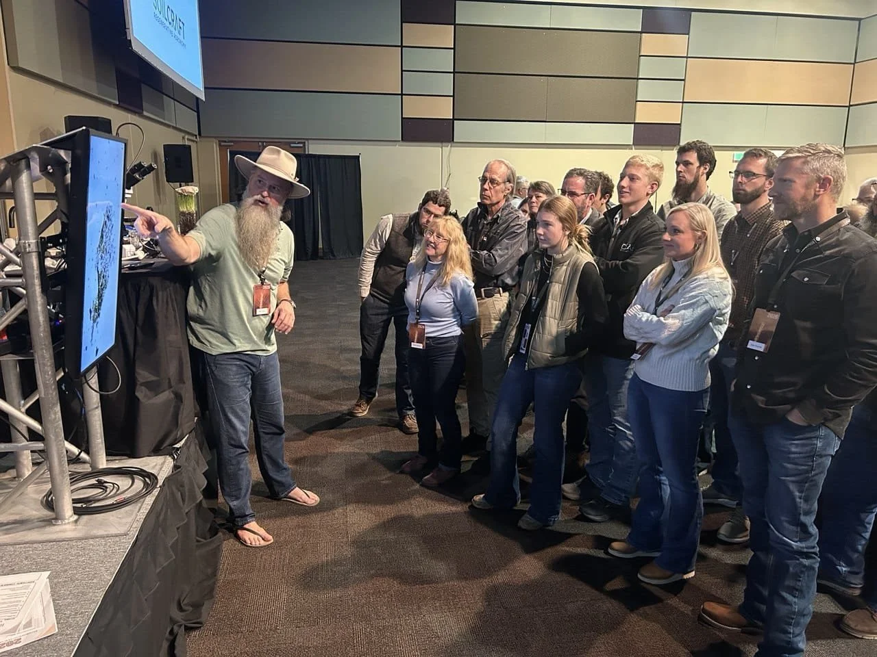 A man with a long beard, wearing a hat, green shirt, and sandals, is talking to a group at a conference. The group includes men and women attentively listening.