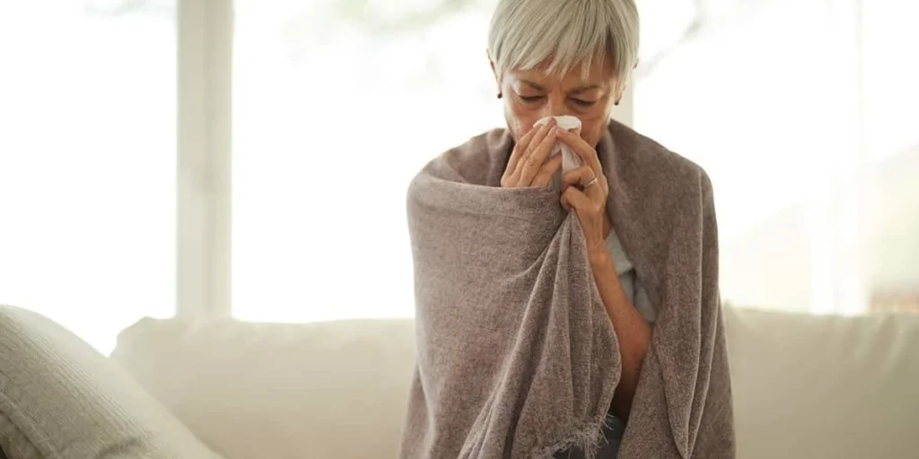 An elderly woman with short gray hair, wrapped in a gray blanket, is holding a tissue to her nose, appearing to be sick.