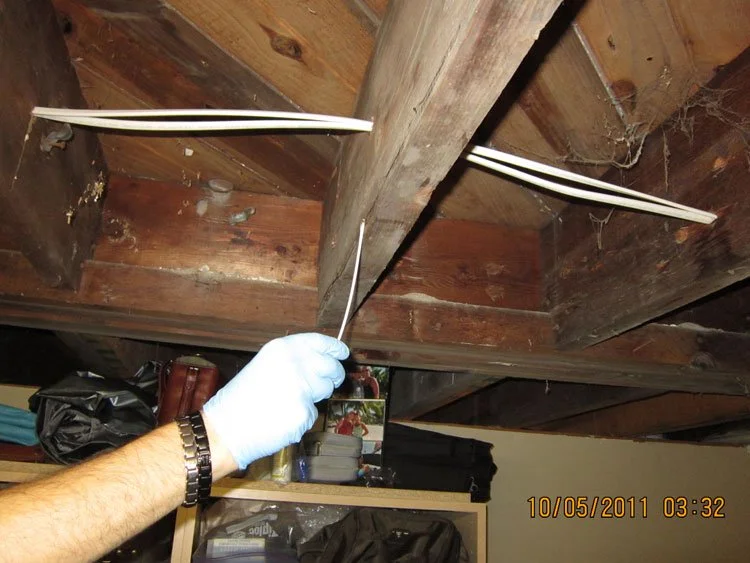 Close-up of a person wearing a white glove and holding a wire or pin, working on electrical wiring or maintenance under a wooden ceiling with exposed beams. There are plastic conduit pipes or tubing running through the ceiling.