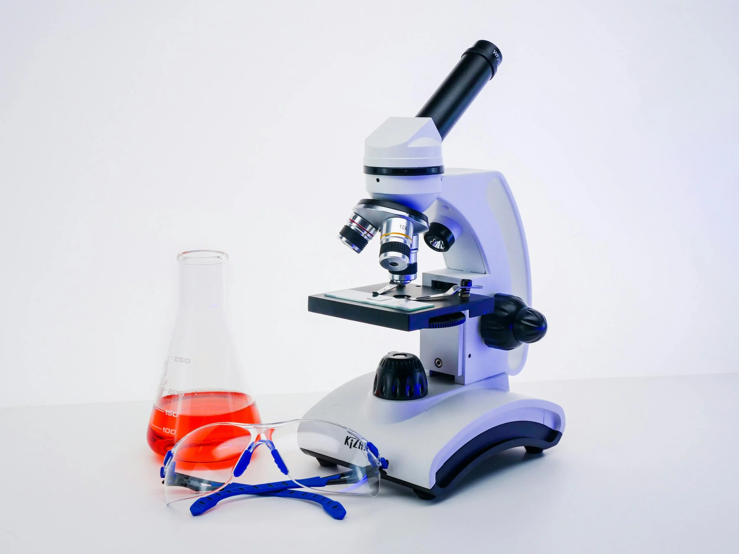 A laboratory microscope with a white and black body, a glass beaker with red liquid, safety glasses, and a blue and white face mask placed on a white surface.