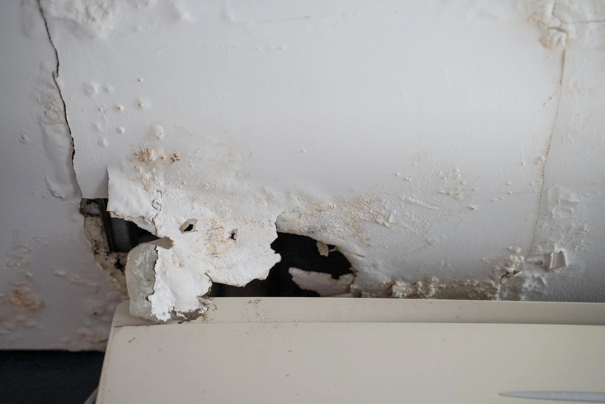 A ceiling with peeling paint and damage showing exposed black wiring and a small section of a white piece of furniture at the bottom.