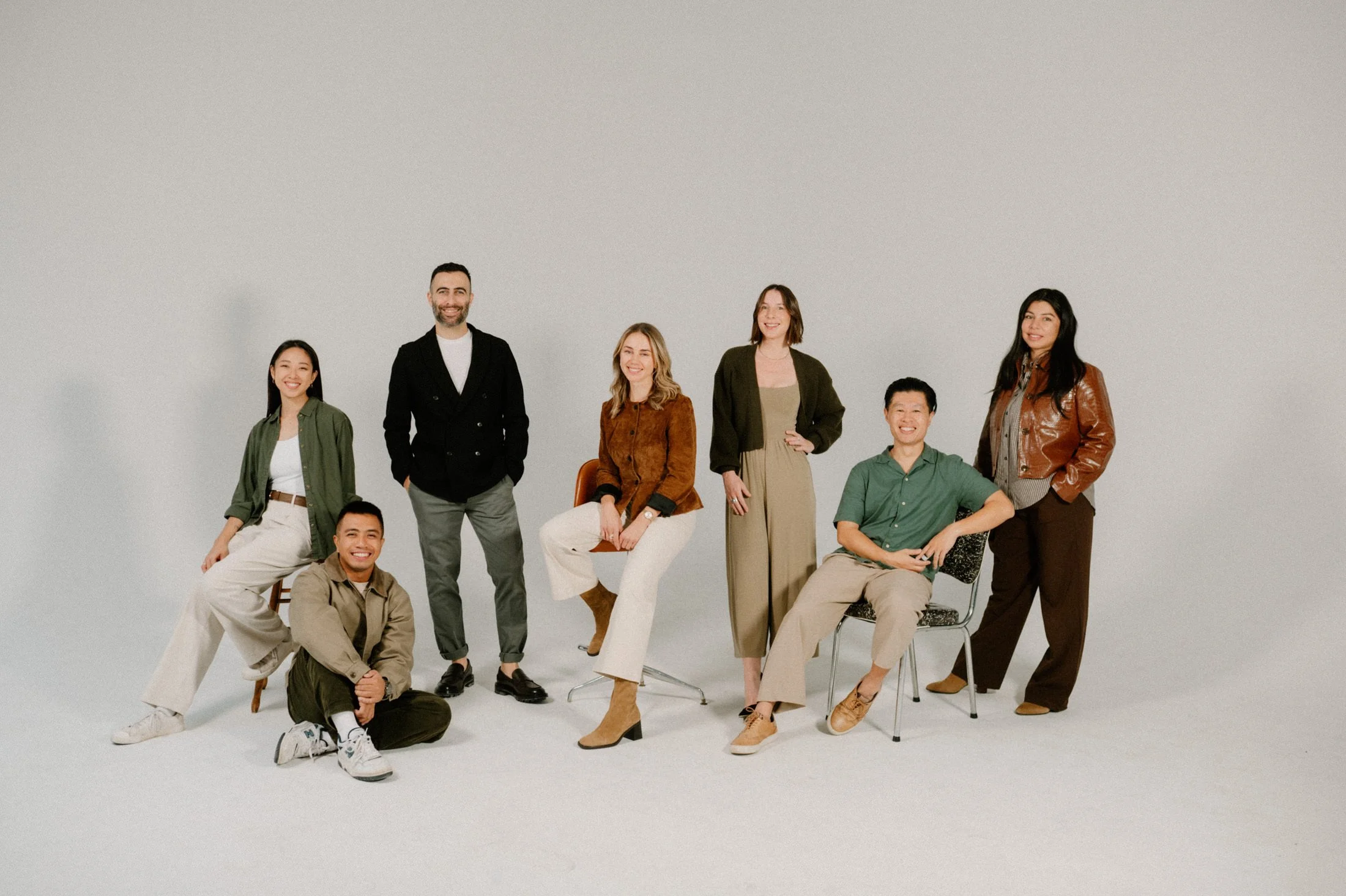 Group of seven diverse people smiling and posing together in a studio against a plain light gray background.