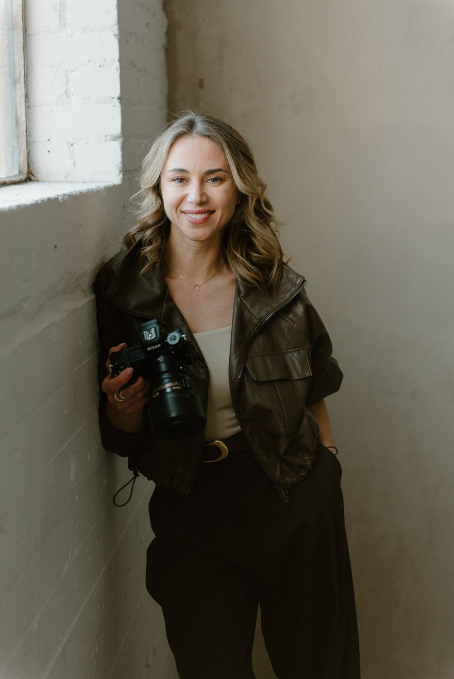 A young woman with wavy blonde hair, smiling, standing in a corner by a window with natural light. She is wearing a dark leather jacket, a light top, and black pants, holding a professional camera in her right hand.