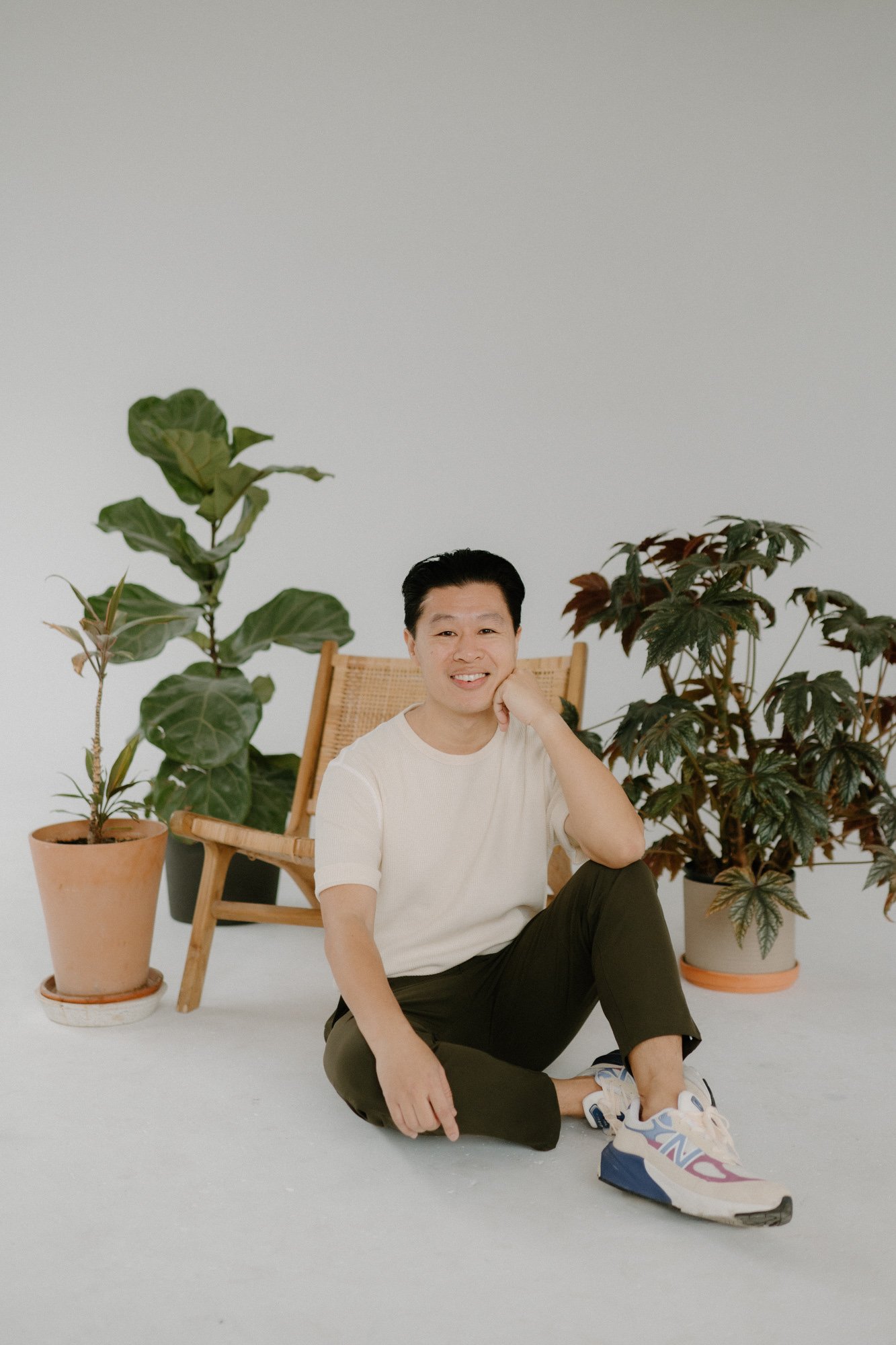 A person sitting on the floor surrounded by potted plants, smiling with hand resting on cheek, in a minimalistic indoor setting.