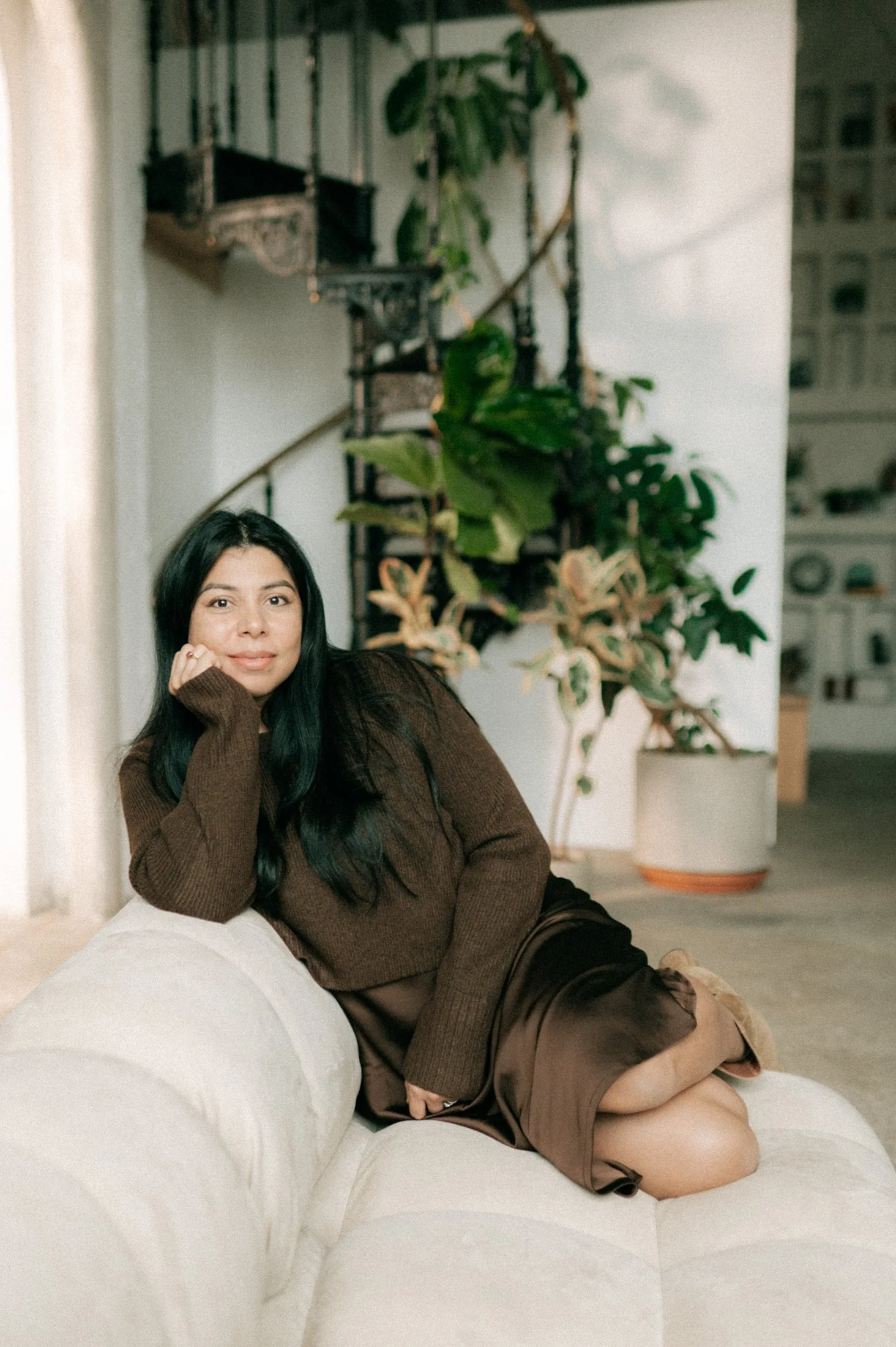 Woman lying on a white couch with her head resting on her hand, wearing a brown sweater and silk skirt, with large green plants and a staircase in the background.