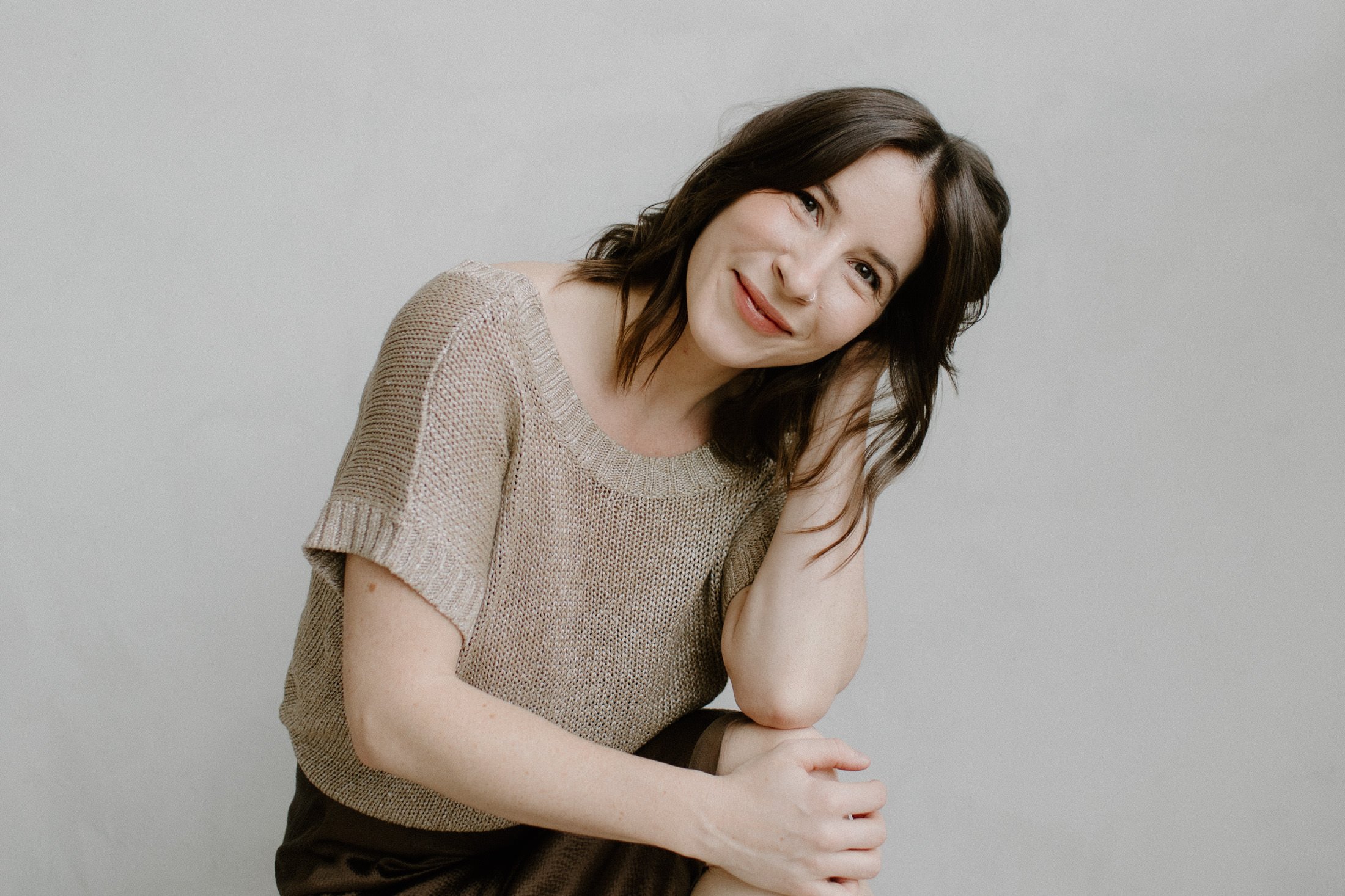 A woman with shoulder-length dark brown hair, smiling and slightly tilting her head, wearing a beige knit short-sleeve top against a plain light gray background.