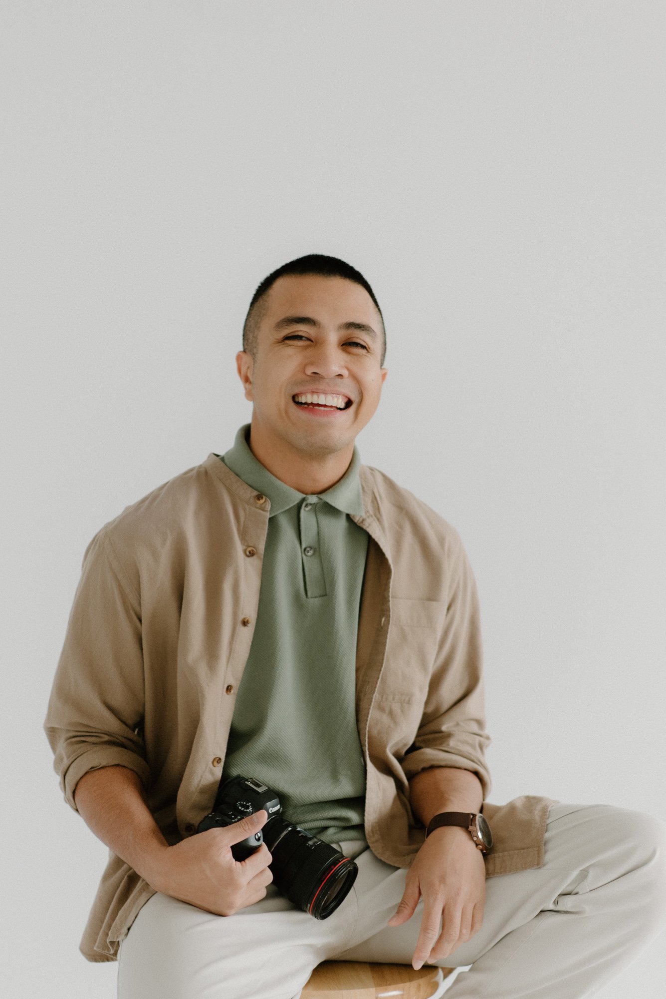 A smiling man sitting on a stool, holding a camera, wearing a beige jacket over a green polo shirt, with light-colored pants, against a plain white background.