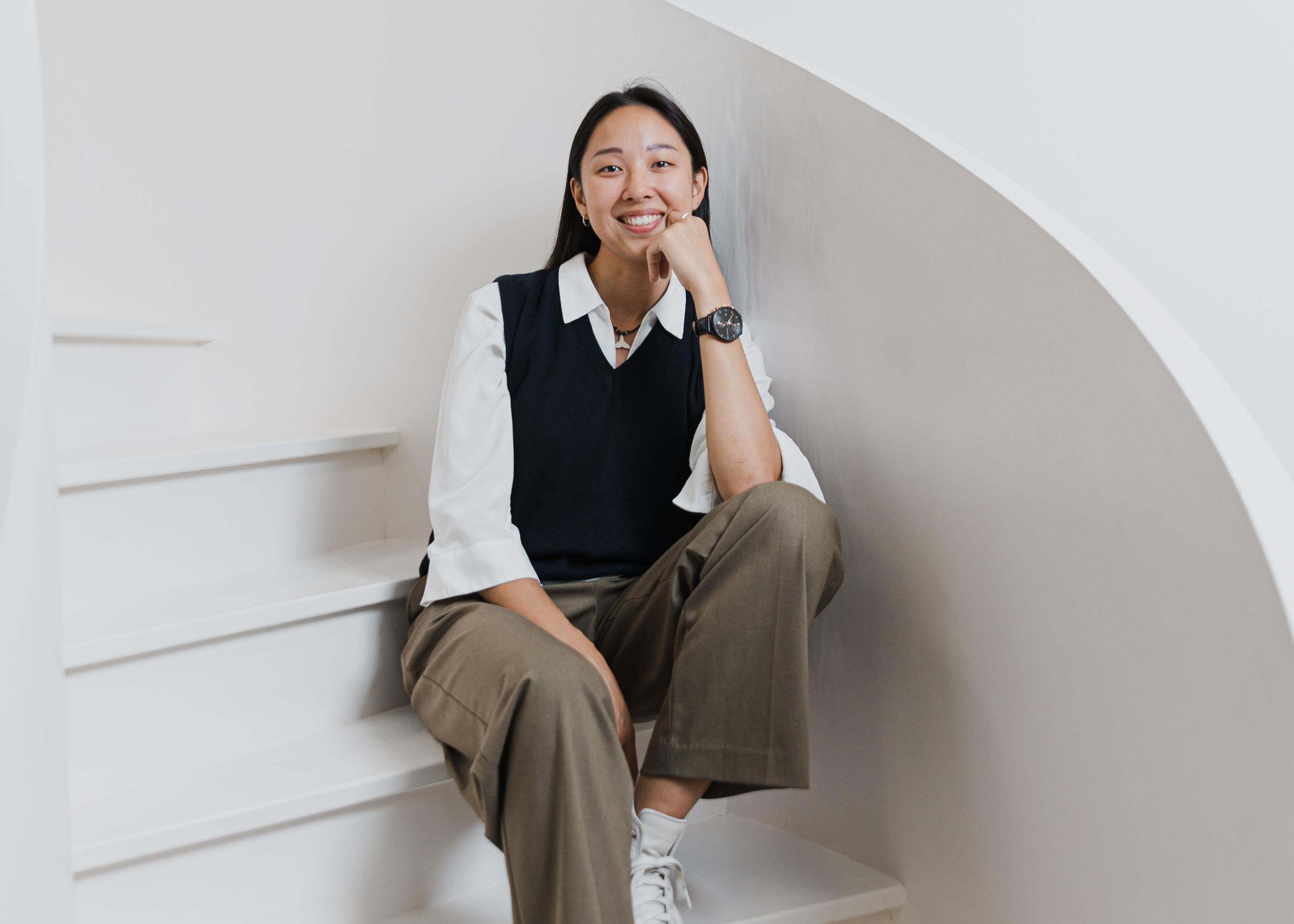 A woman sitting on white stairs, smiling, wearing a white blouse with ruffled sleeves, a dark sleeveless sweater, brown pants, white sneakers, and a black wristwatch.