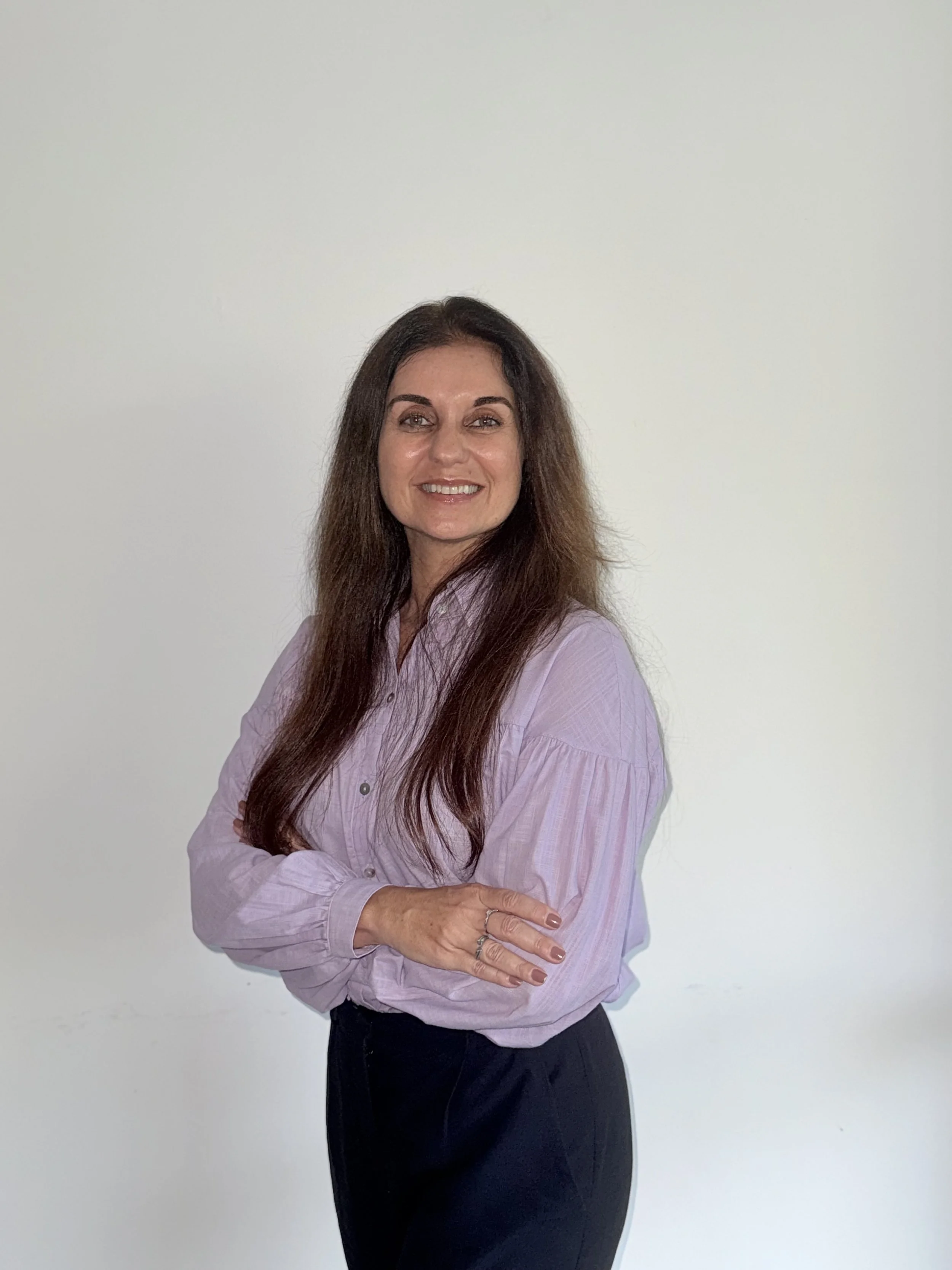 A woman with long brown hair, wearing a light purple blouse and dark pants, standing against a plain white wall. She is smiling and has her arms crossed.