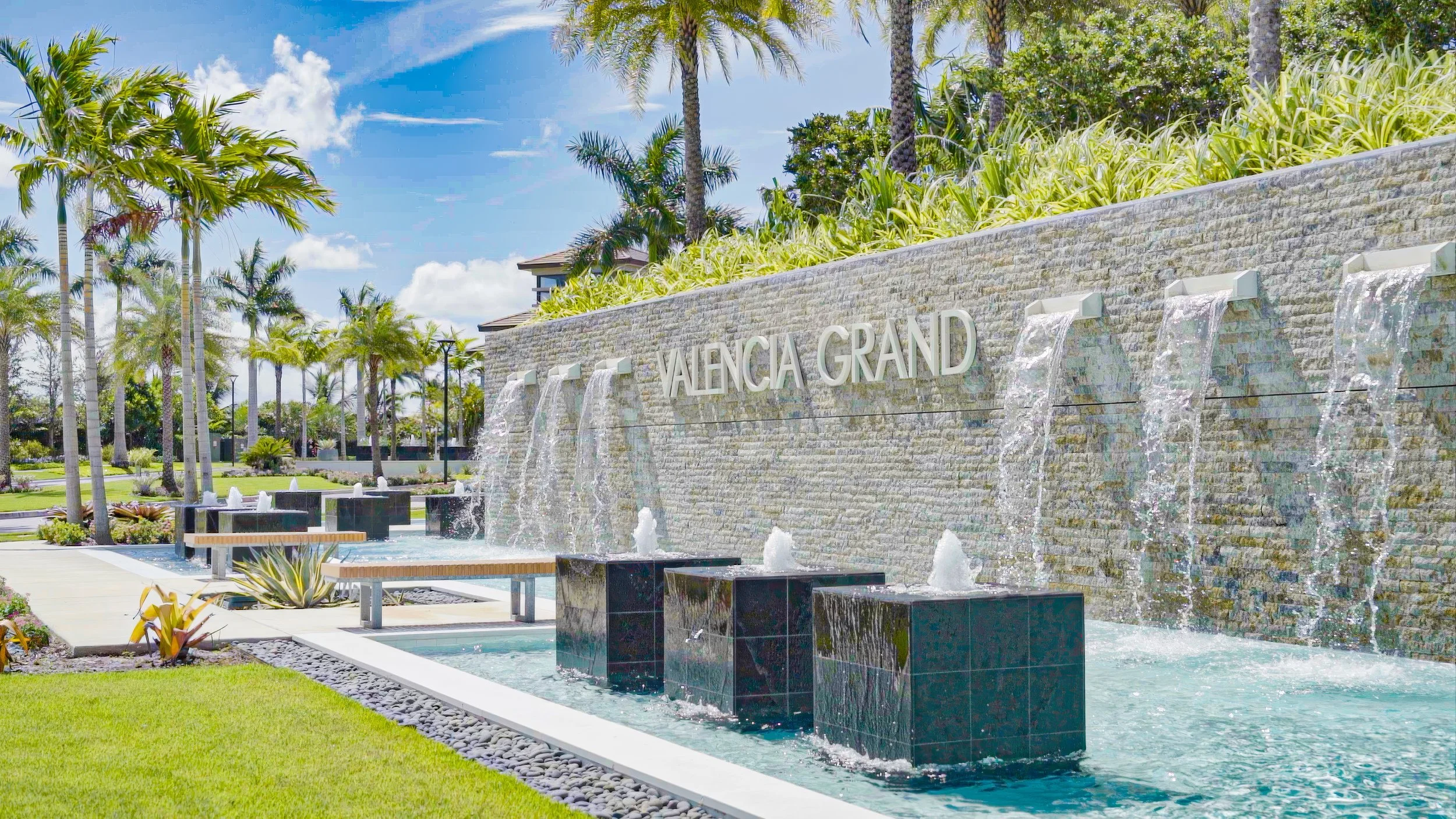 Valencia Grand hotel entrance with a large stone fountain feature, multiple waterfalls, lush tropical landscaping, and palm trees under a bright blue sky.