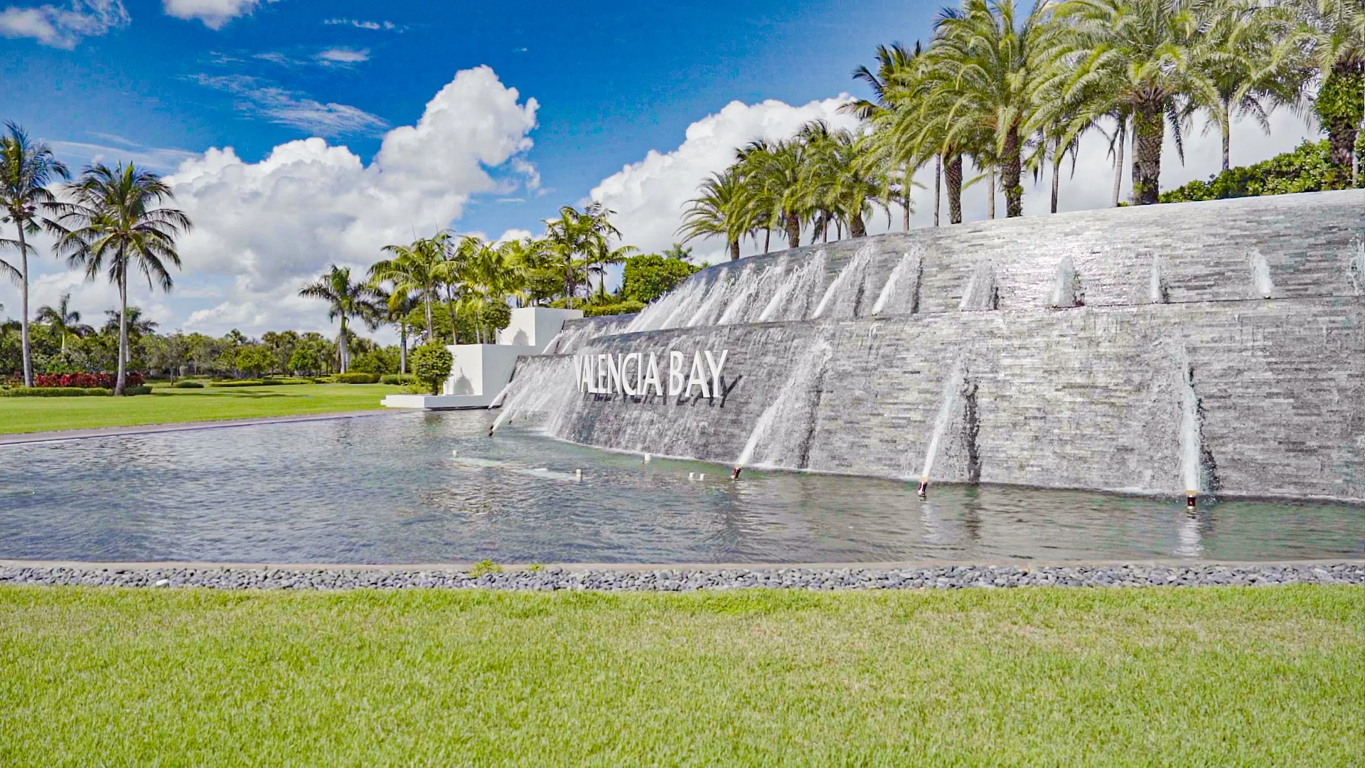A water fountain with the sign "Valencia Bay" surrounded by palm trees and green grass in a sunny outdoor area.