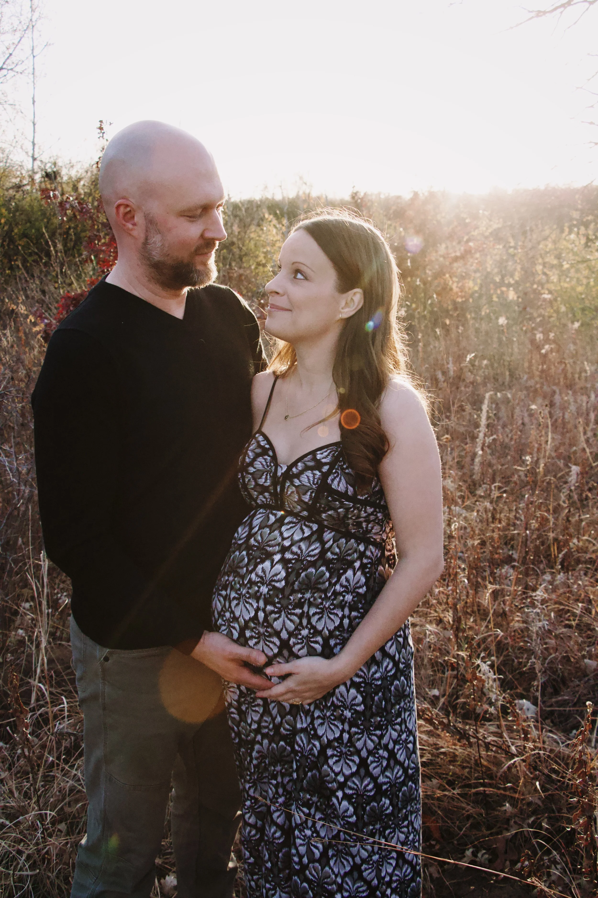 Couple embracing outdoors at sunset, with the woman cradling her pregnant belly.