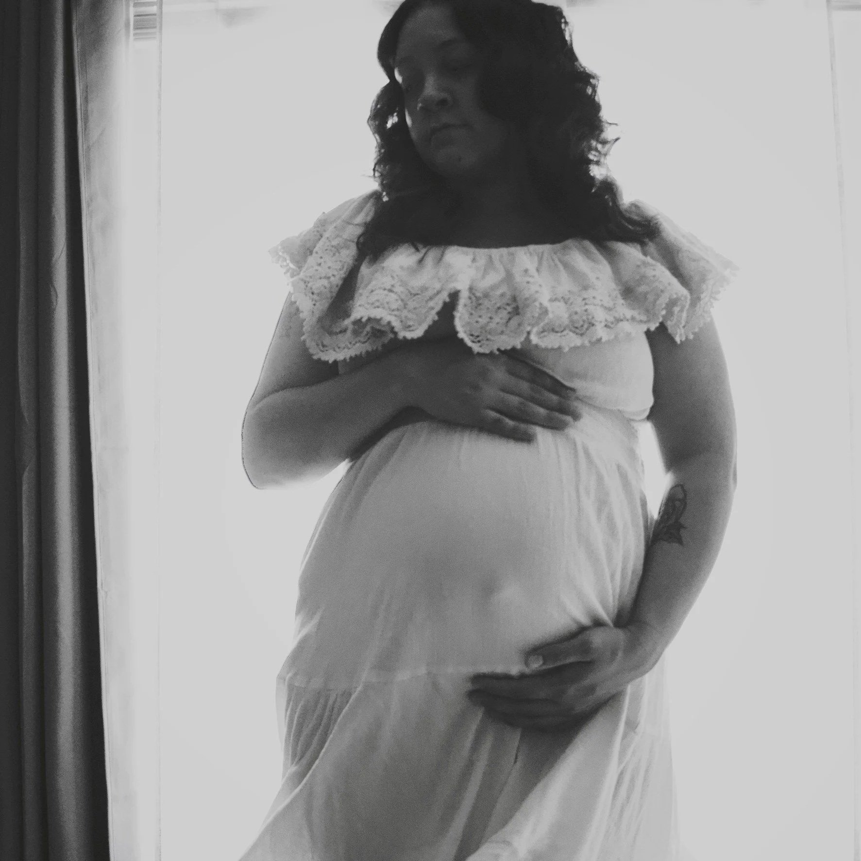 Black-and-white portrait of a pregnant woman in a vintage lace-collar dress cradling her belly by a window.