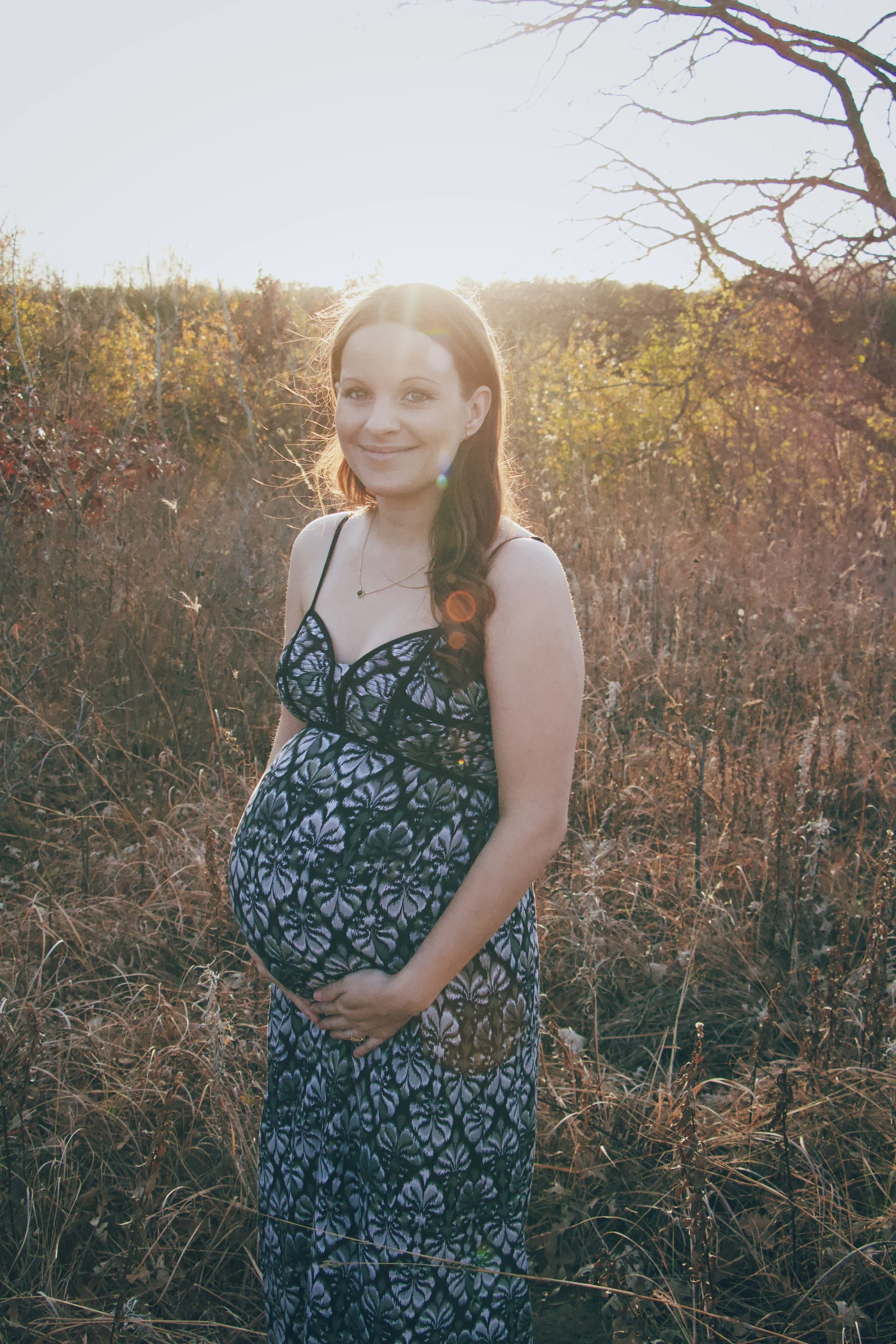 Pregnant woman in a patterned dress standing outdoors in a sunlit autumn field