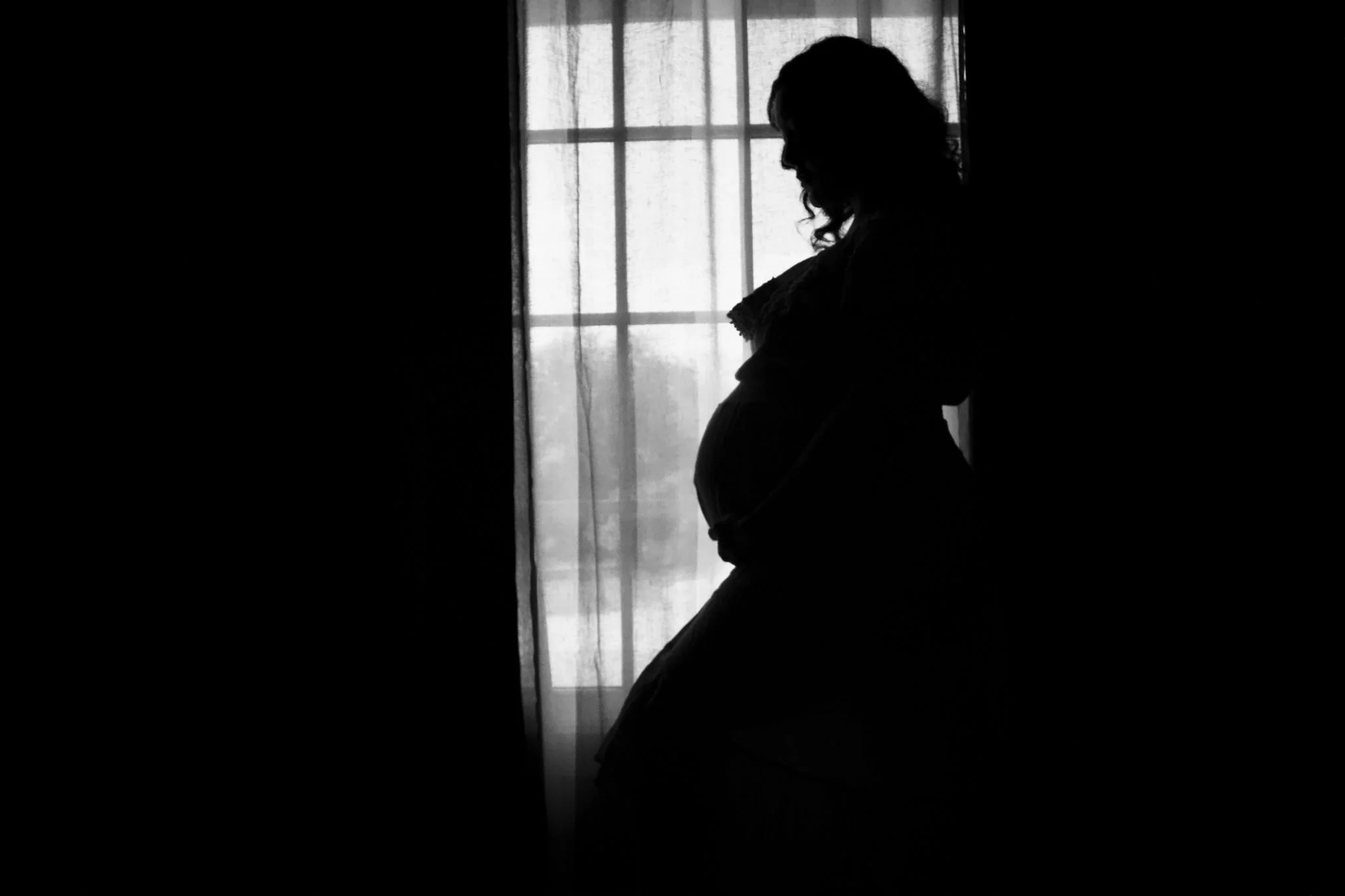Black-and-white silhouette of a pregnant woman standing in front of a curtained window