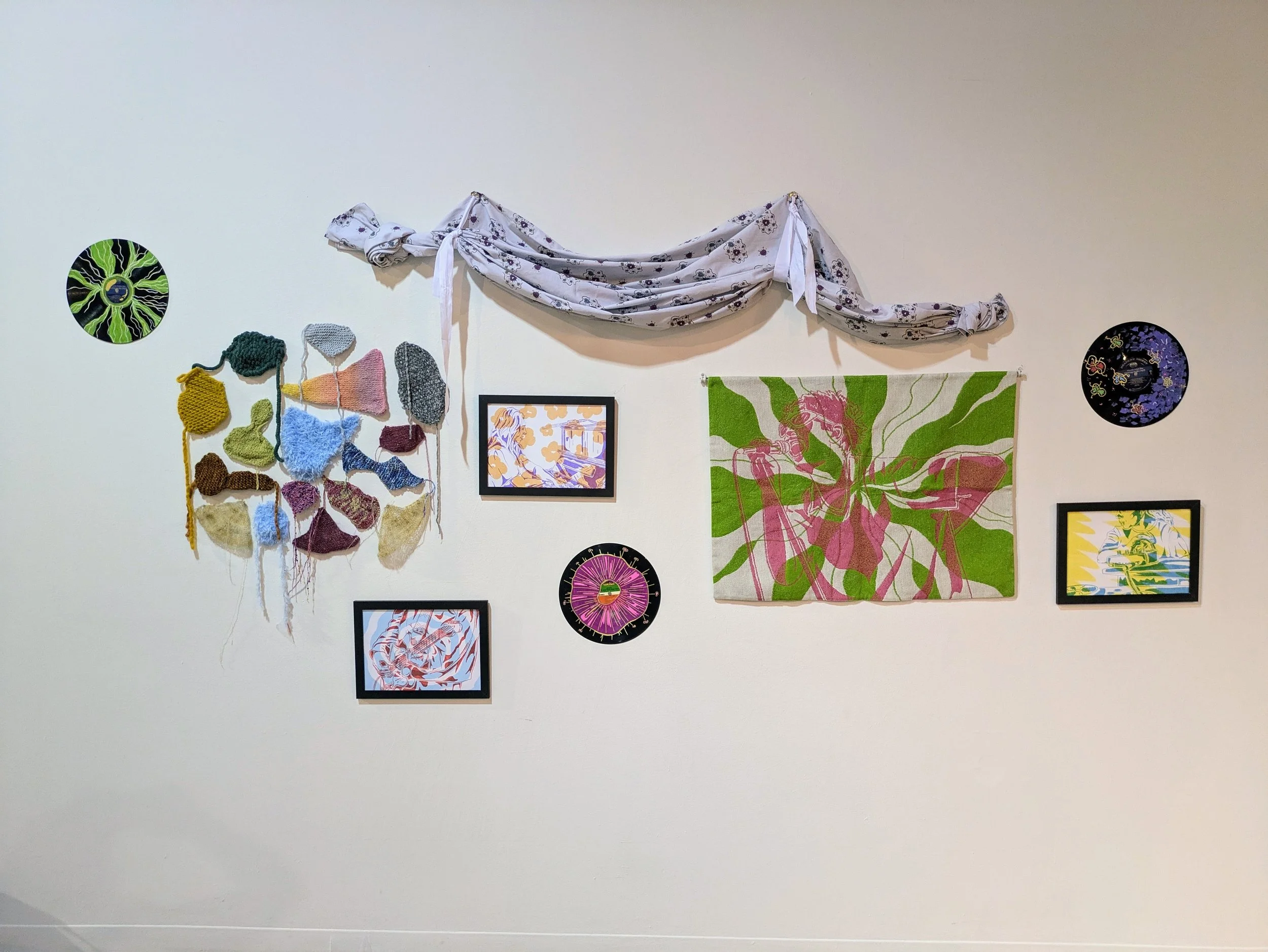 Wall decorated with hanging knitted and crocheted art, framed pictures, circular decorative pieces, and a fabric drape, creating an eclectic, artistic display.